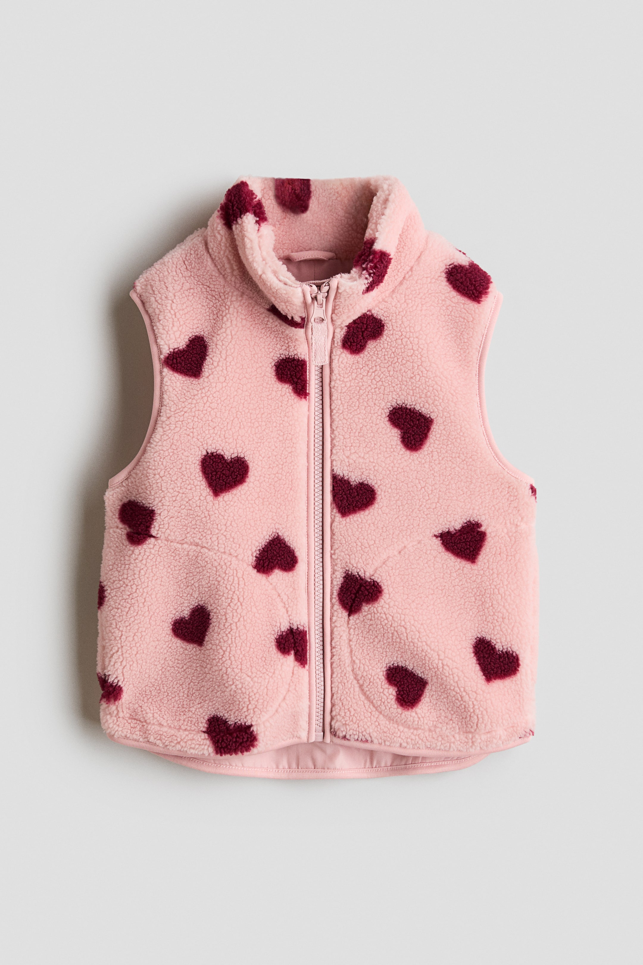 View larger image: A pink sherpa vest displaying a scattered heart pattern in burgundy. It features a full-length front zipper, a stand-up collar, and two integrated front pockets.