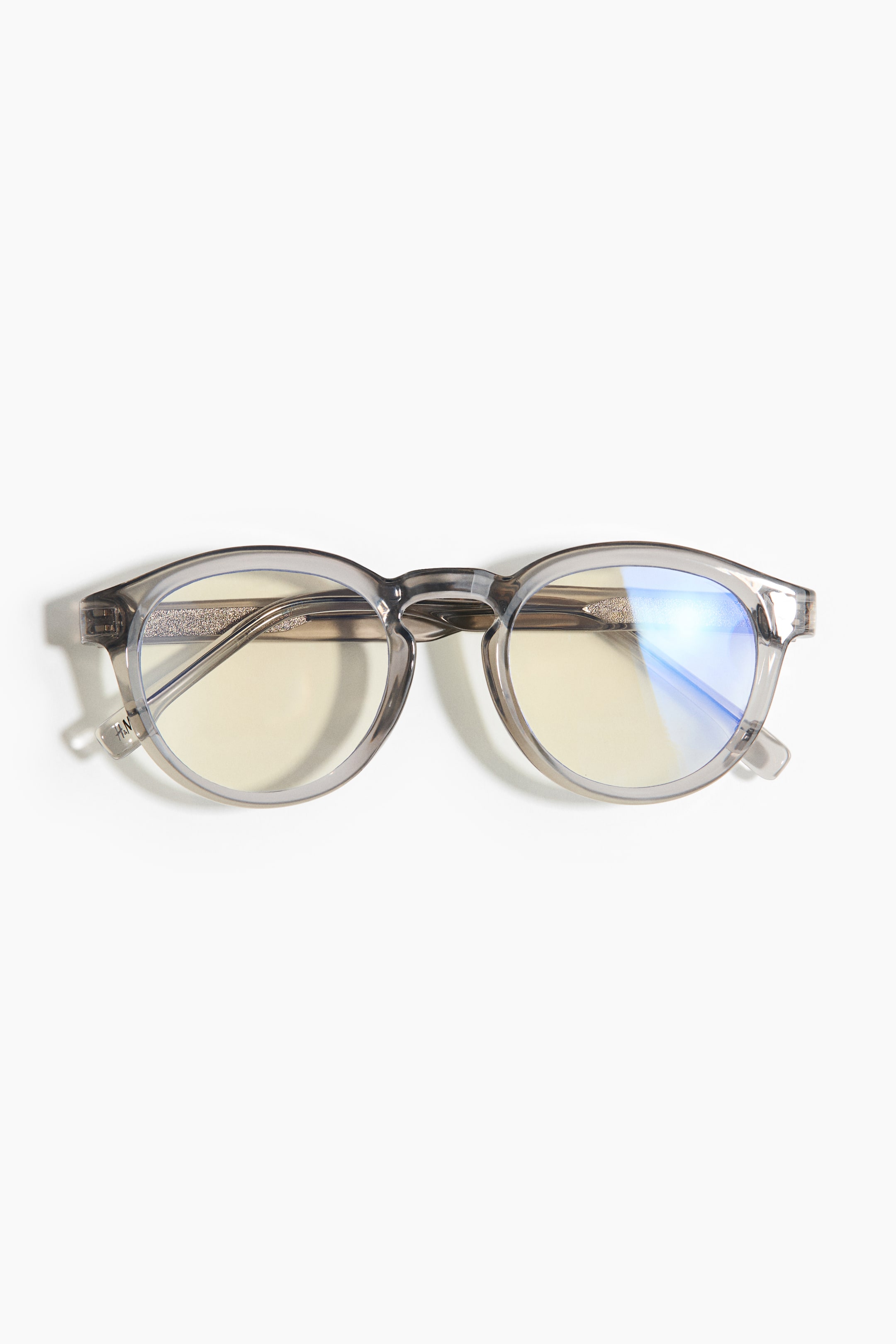 View larger image: A pair of eyeglasses is shown in a front view, featuring a translucent gray frame and rounded lenses. The lenses exhibit a yellowish tint, with the right lens reflecting a blue light. Subtle textured detailing is visible on the inner temples.