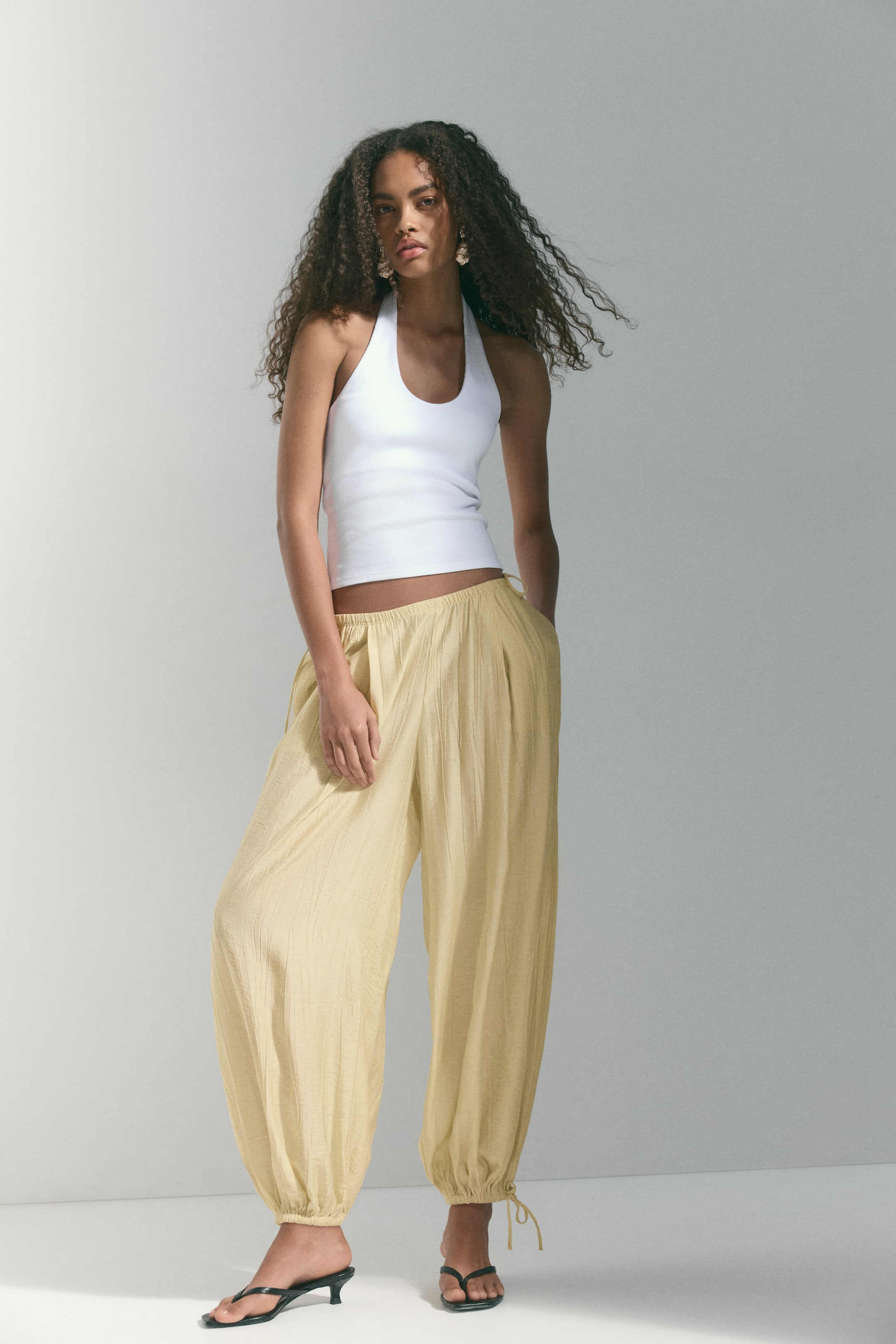 View larger image: A white halter neck top boasts a U-neckline and a fitted, slightly cropped silhouette, paired with light yellow crinkled wide-leg trousers that gather at the ankles with visible ties.