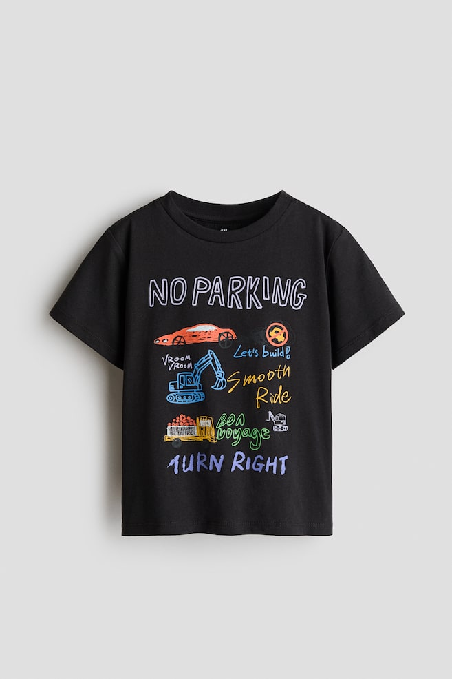 黒の半袖Tシャツの前面にカラフルなグラフィックデザインが施されています。クルーネックで、デザインにはオレンジ色の車、青いショベルカー、黄色のトラックのアウトラインが含まれています。また、「NO PARKING」、「Let's build!」、「Smooth Ride」、「Bon voyage」、「TURN RIGHT」といったテキストが様々な鮮やかな色で描かれています。 - 1