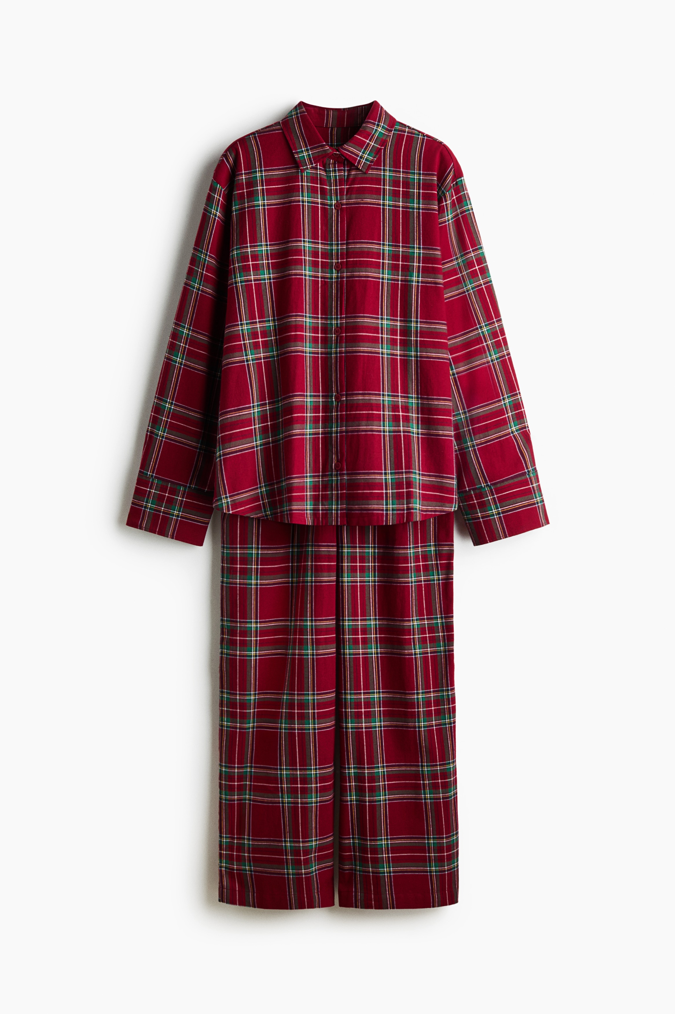 A red plaid flannel pyjama set is worn by the model, featuring a long-sleeved, collared button-down shirt and matching wide-leg pants. The shirt is left partially unbuttoned at the lower hem, revealing the elasticated drawstring waistband of the pants.