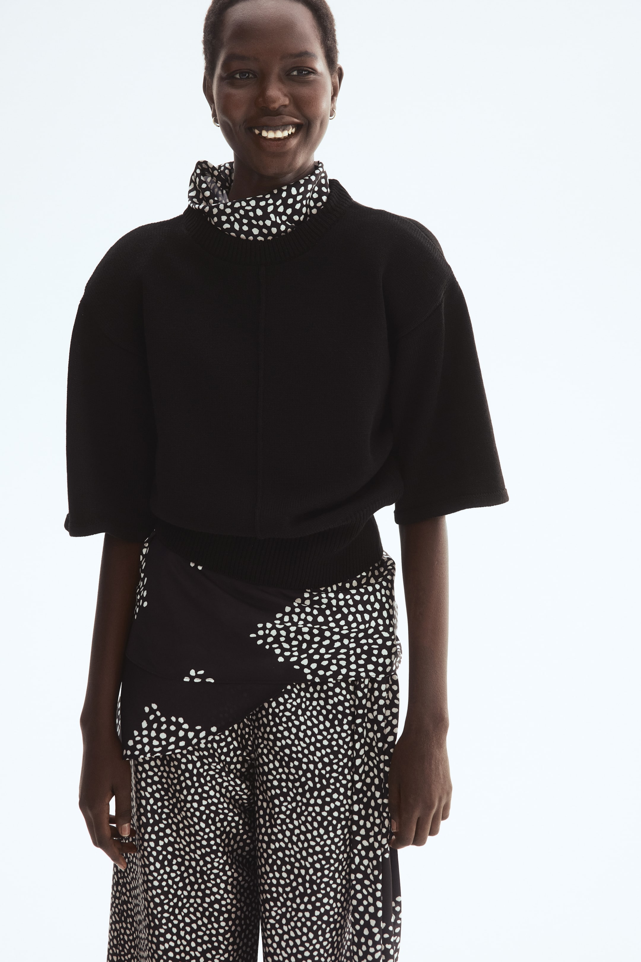 Ingrandisci l'immagine: Un top lavorato a maglia, caratterizzato da una cucitura collegata lungo la parte anteriore, spalle scese e maniche corte, è indossato dalla modella. È visibile una costina attorno al collo, ai polsini e all'orlo, e un indumento a collo alto con motivo è stratificato sotto il top.