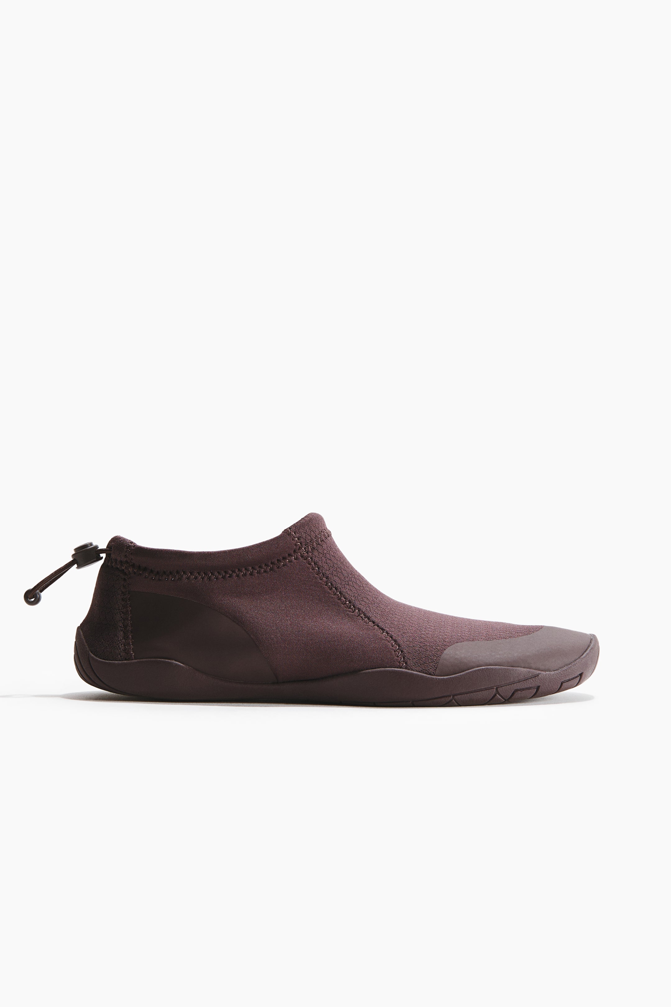 Agrandir l'image: Une chaussure marron foncé à enfiler présente son profil latéral. Elle est dotée d'une tige souple et flexible avec des coutures apparentes, un embout renforcé en caoutchouc et un cordon noir avec un arrêt à bascule au talon. La chaussure repose sur une semelle texturée, profil bas, de la même couleur marron foncé.