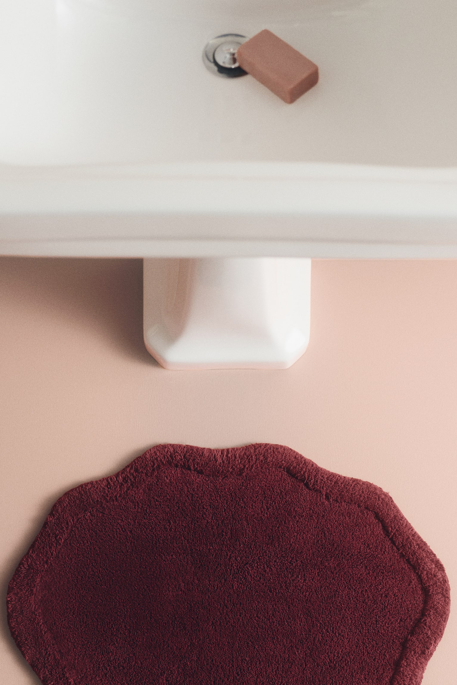An overhead view shows a burgundy tufted cotton bath mat featuring a scalloped, art deco-inspired silhouette and a thick raised border. The mat is lying flat on a light pink floor, positioned directly in front of the base of a white pedestal sink. - 1