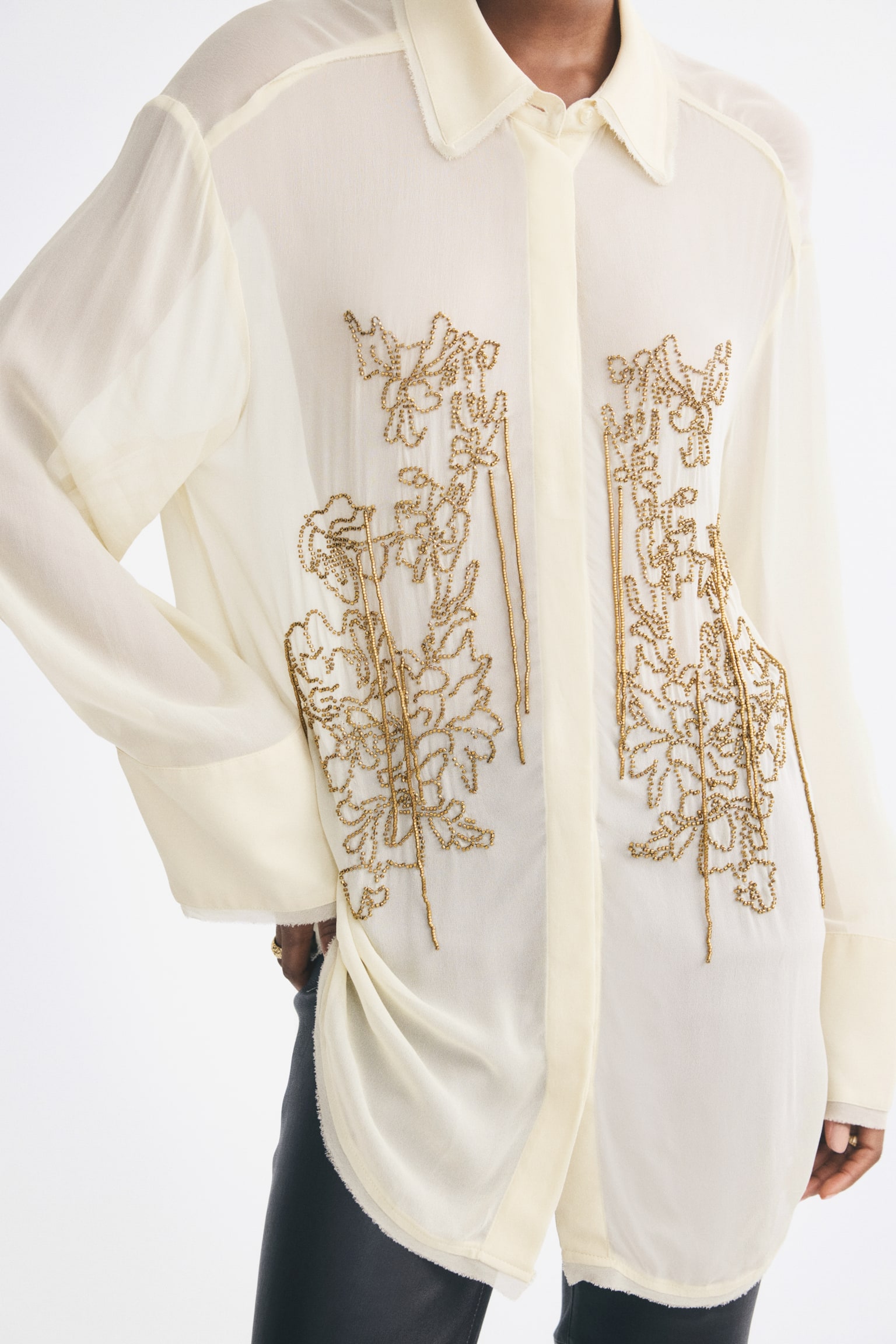 A semi-sheer, off-white beaded viscose shirt is worn by the model, featuring intricate gold floral beadwork with hanging strands on the front panels. The long-sleeved shirt has a classic collar and wide cuffs, and is styled with dark trousers. - 1