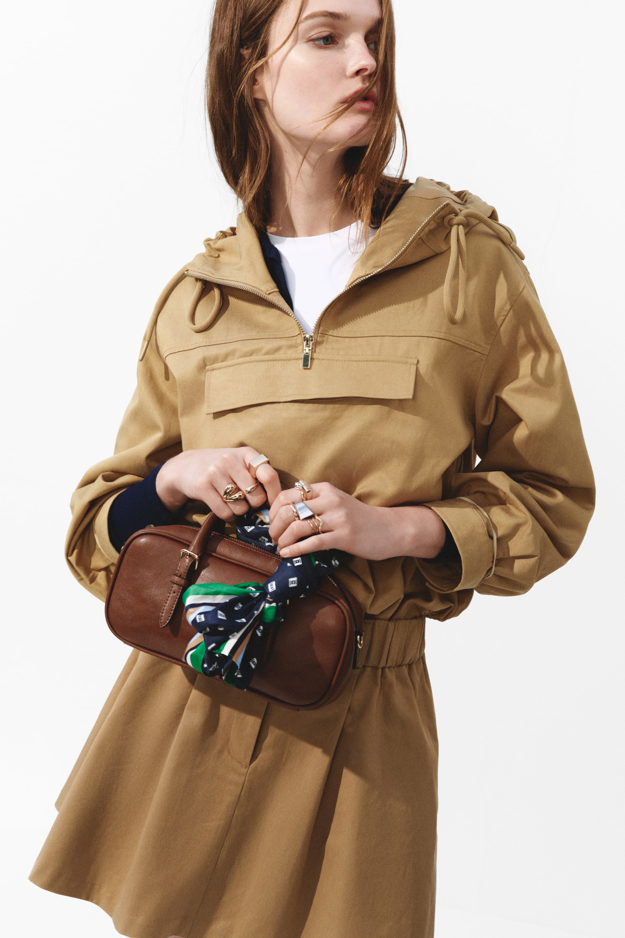 View larger image: A tan anorak-style one-piece garment features a half-zip closure with a gold-toned zip, a drawstring hood, and a large front flap pocket. It presents long sleeves with buttoned cuffs, a cinched waist creating a blouson effect, and a full, pleated lower section, while worn over a white t-shirt and a navy layer.