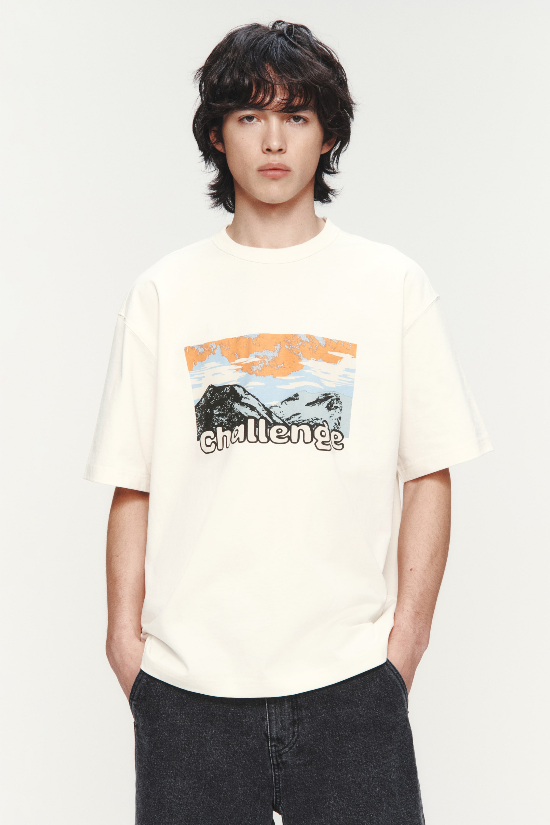 Xem ảnh lớn hơn: Áo thun ngắn tay màu trắng ngà với cổ tròn và dáng rộng, nhìn từ phía trước. Áo có hình in phía trước mô tả những ngọn núi tối dưới bầu trời với mây màu cam và xanh, cùng chữ "challenge" màu trắng nằm chồng lên những ngọn núi.
