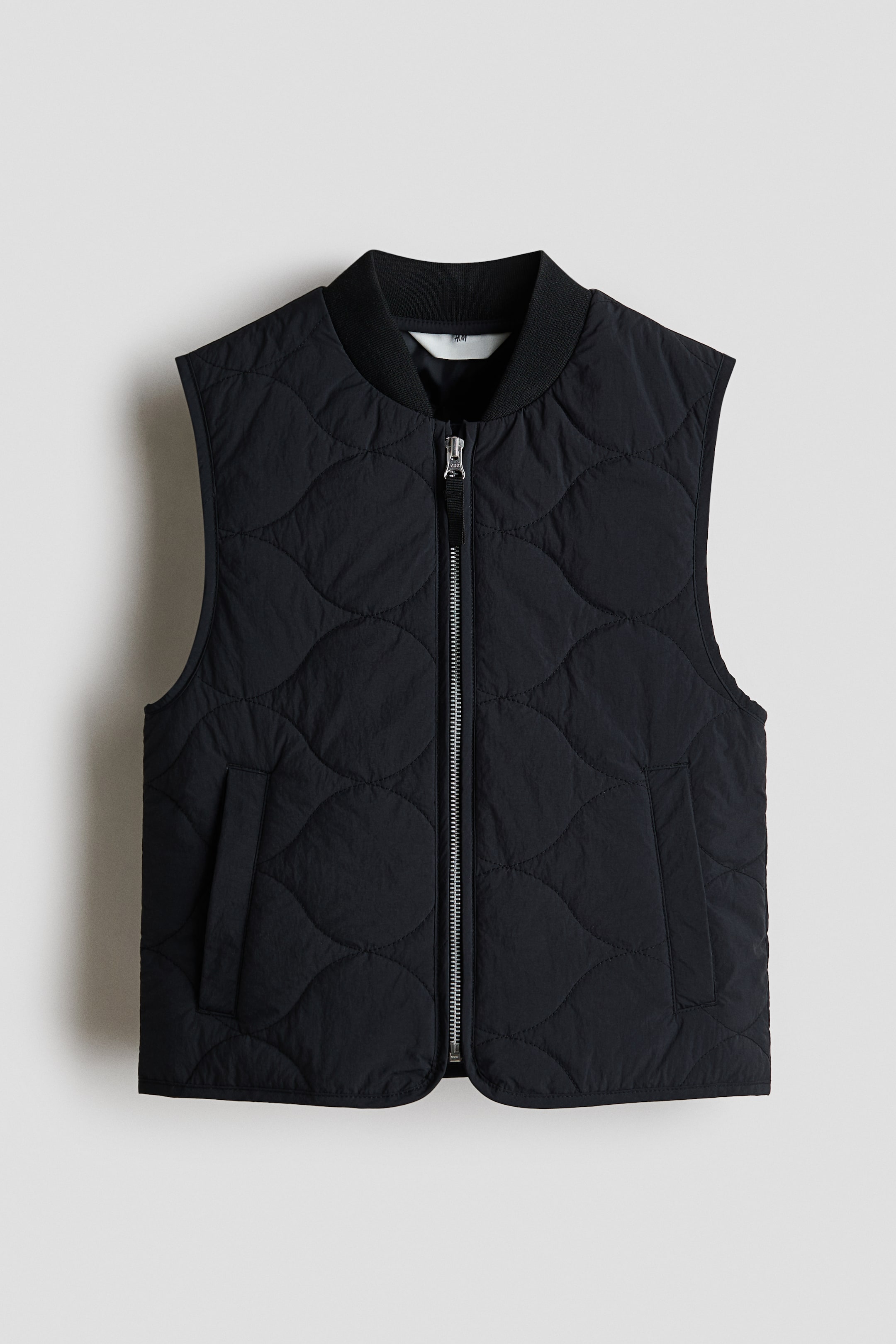Ampliar la imagen: Un chaleco negro se muestra de frente, presentando un tejido de nylon acolchado con un patrón ondulado. Está diseñado con un cuello de banda acanalada, un cierre central con cremallera y protector de barbilla antirozaduras, dos bolsillos laterales de ojal y un dobladillo de corte recto.