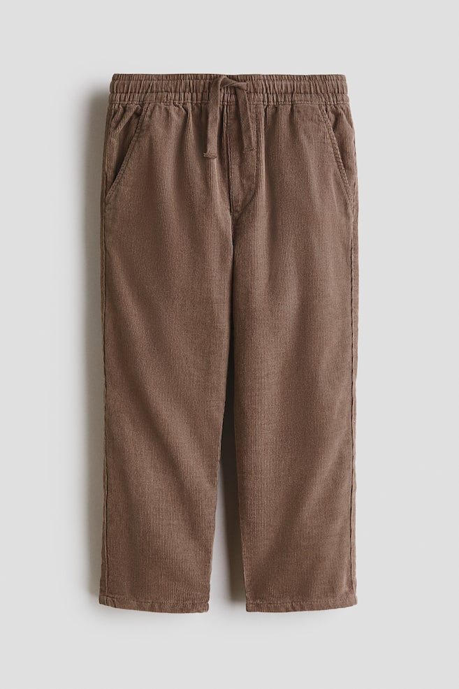 Soft brown corduroy trousers are presented from the front, featuring an elasticated drawstring waist, a fake fly, and side slash pockets, with a loose fit through the hip and thigh and legs gently tapering towards the hem. - 1
