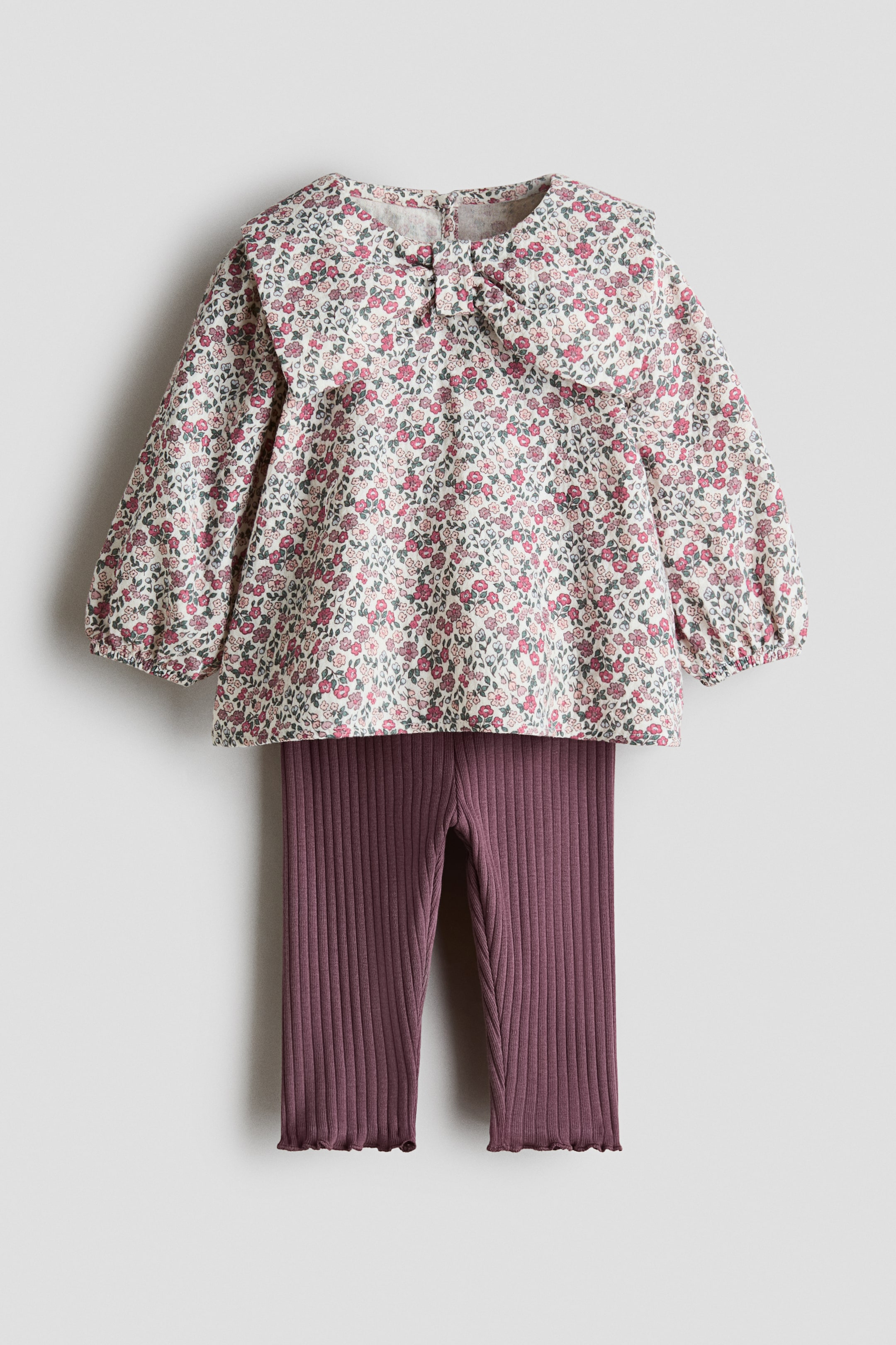 View larger image: A front-facing set is featuring a floral blouse with an oversized, bow-shaped collar and dark purple ribbed leggings. The blouse is displaying a small button at the center of its collar and long sleeves gathering at elasticated cuffs, while the leggings are showcasing overlocked hems at the ankles.