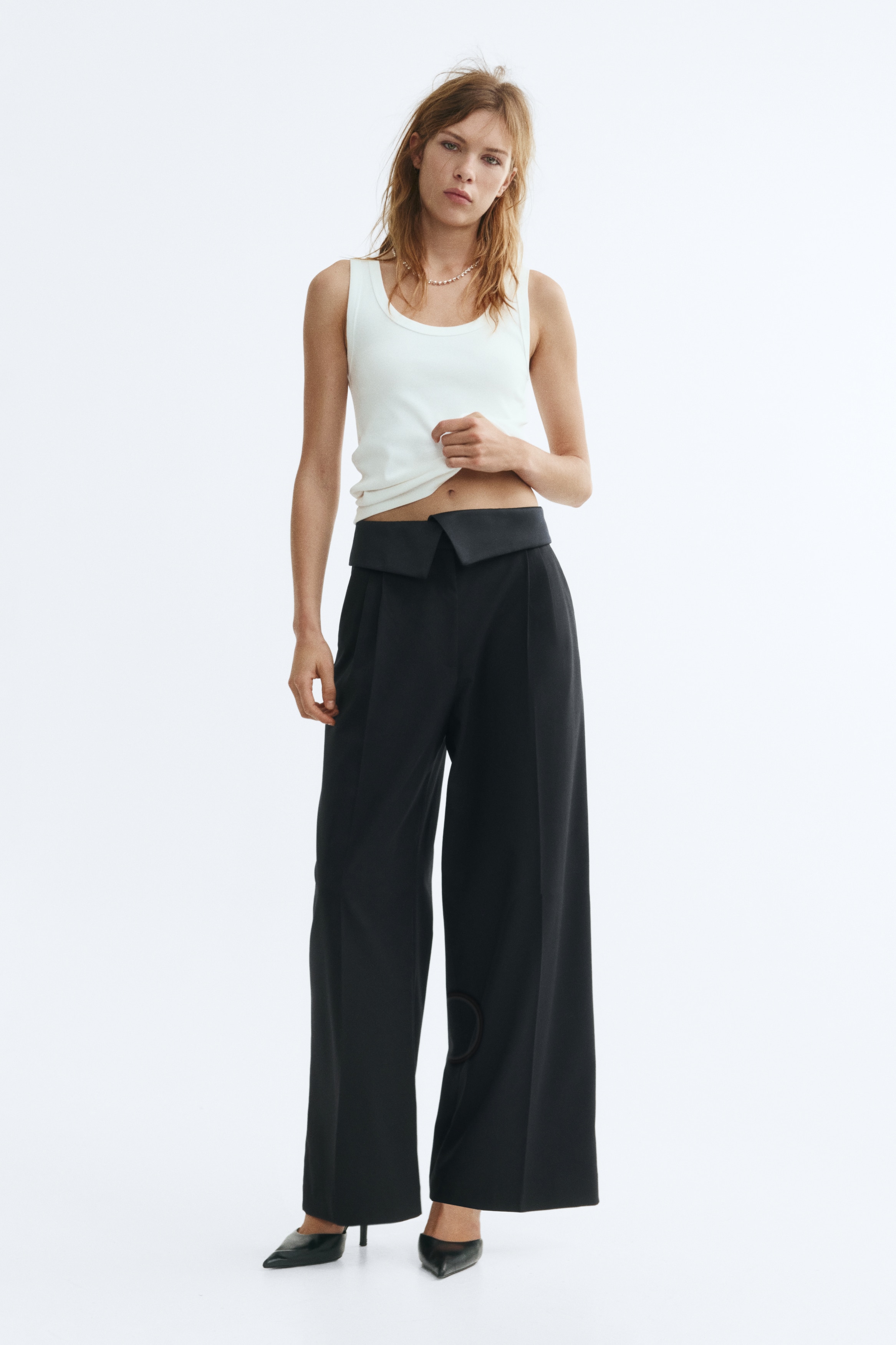 Black loose-fit trousers in woven fabric are shown from the front, highlighting a high waist with a wide, overlapping foldover waistband. The trousers feature front pleats, subtle diagonal side pockets, and wide legs with sharp creases running down the centre.