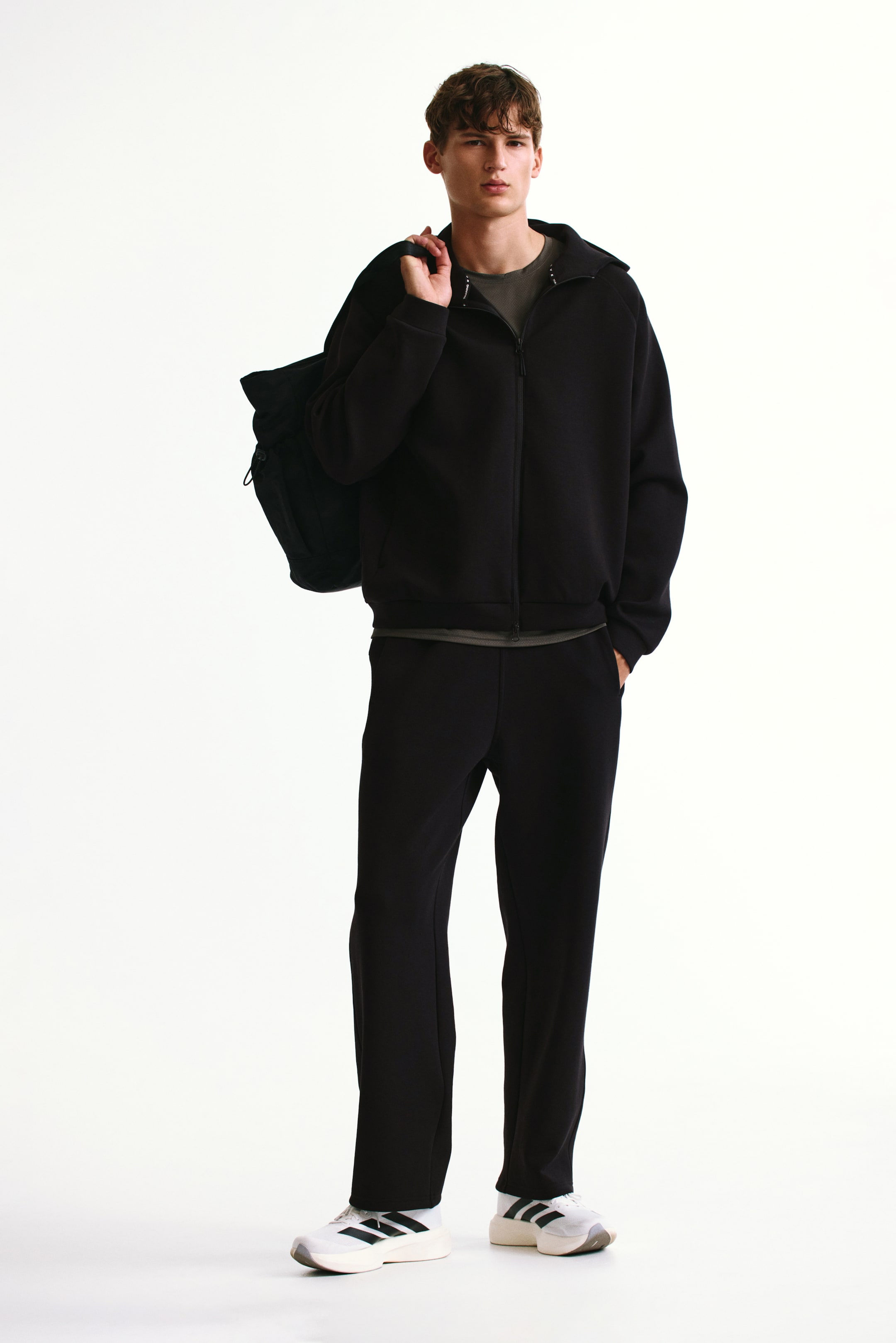 View larger image: A black sports zip-through hoodie, featuring a full-length front zipper and a stand-up collar, is being worn by the model over a dark grey T-shirt. The model is also carrying a black duffel bag on their shoulder, resting against the hoodie.