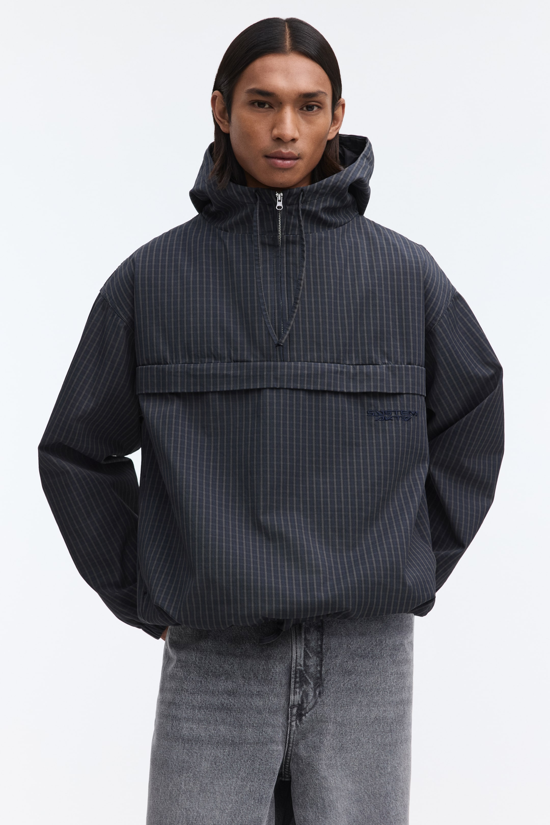 Grotere afbeelding bekijken: Een donkergrijze anorak met capuchon, die recht naar voren is gericht, toont een subtiel krijtstreeppatroon, een halve ritssluiting bij de halslijn en een grote horizontale klepzak over de borst.