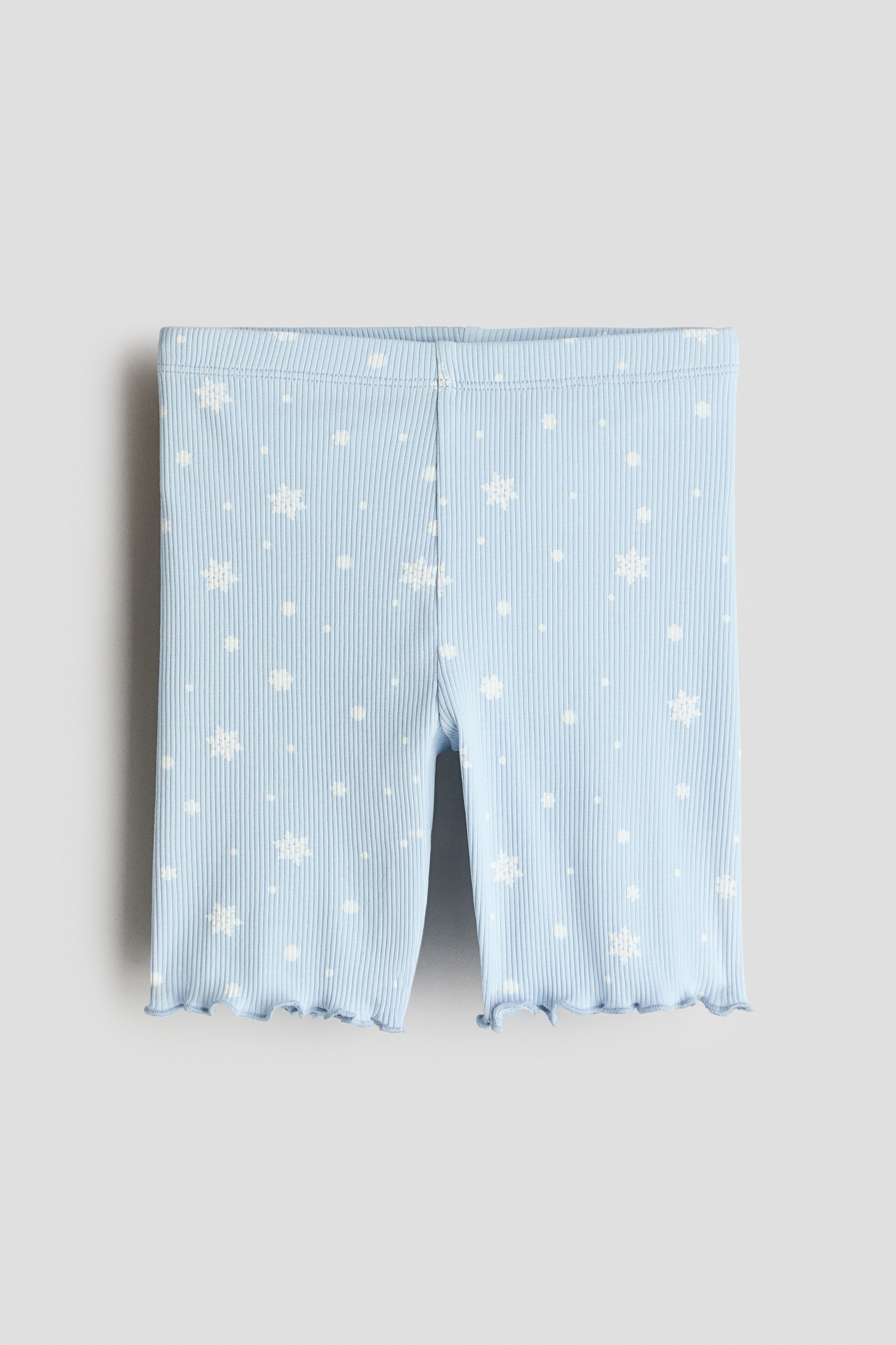Light blue ribbed cotton jersey cycling shorts featuring an all-over white snowflake print and overlocked, wavy hems are being worn by the model. The shorts are styled with a matching light blue sleeveless top with a ruffled collar.