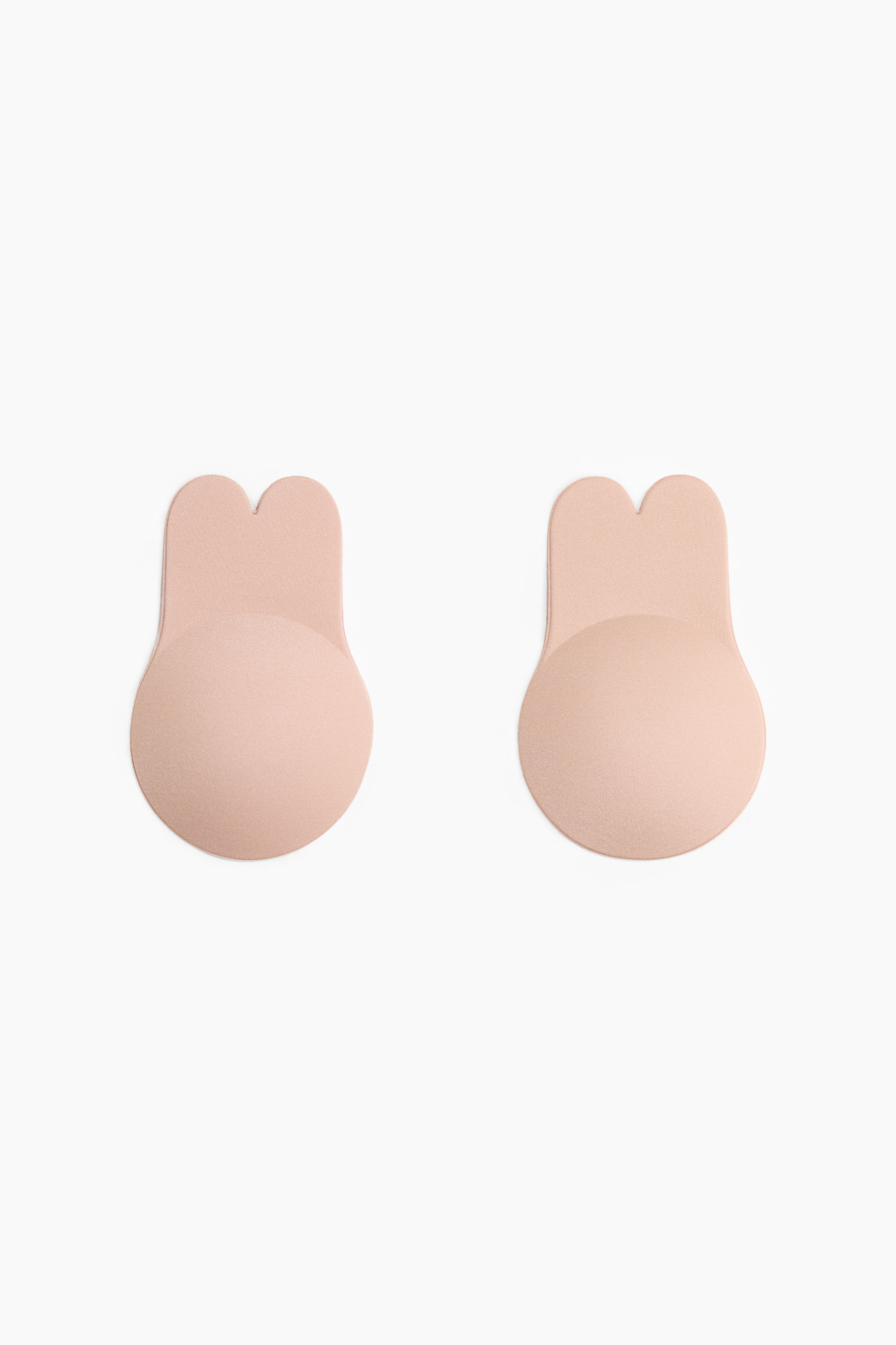 View larger image: A pair of nude-toned nipple covers are laid flat and facing forward. Each cover features a circular base with an upward tab extending, splitting into two rounded lobes at its peak.