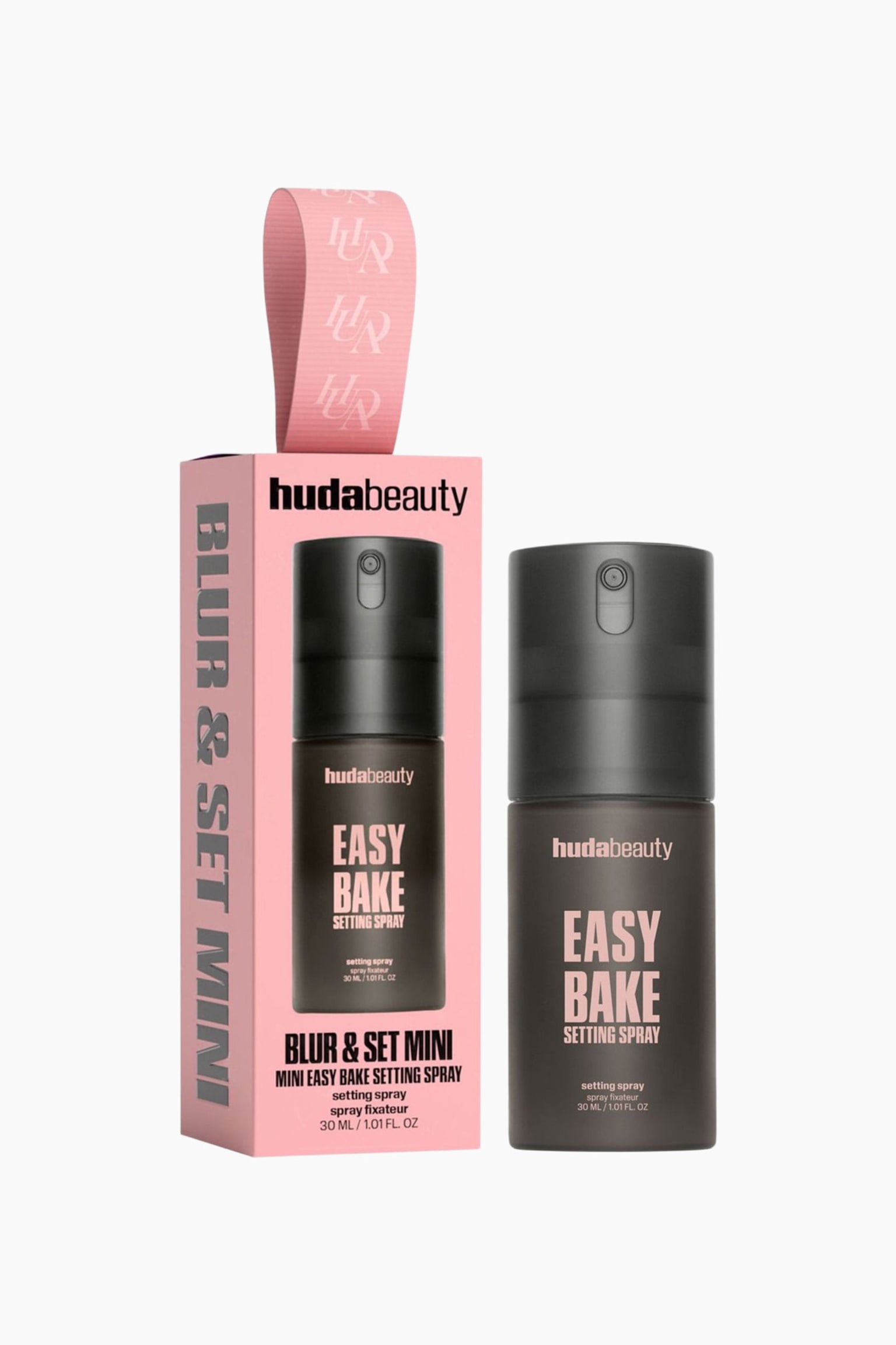 The Huda Beauty Blur & Set Mini Kit is presented in a pink rectangular box with a patterned ribbon loop on the top, alongside a mini Easy Bake Setting Spray bottle. The dark, cylindrical bottle stands upright, showcasing pink text that reads "hudabeauty EASY BAKE SETTING SPRAY", matching the product graphic displayed on the front of the packaging.