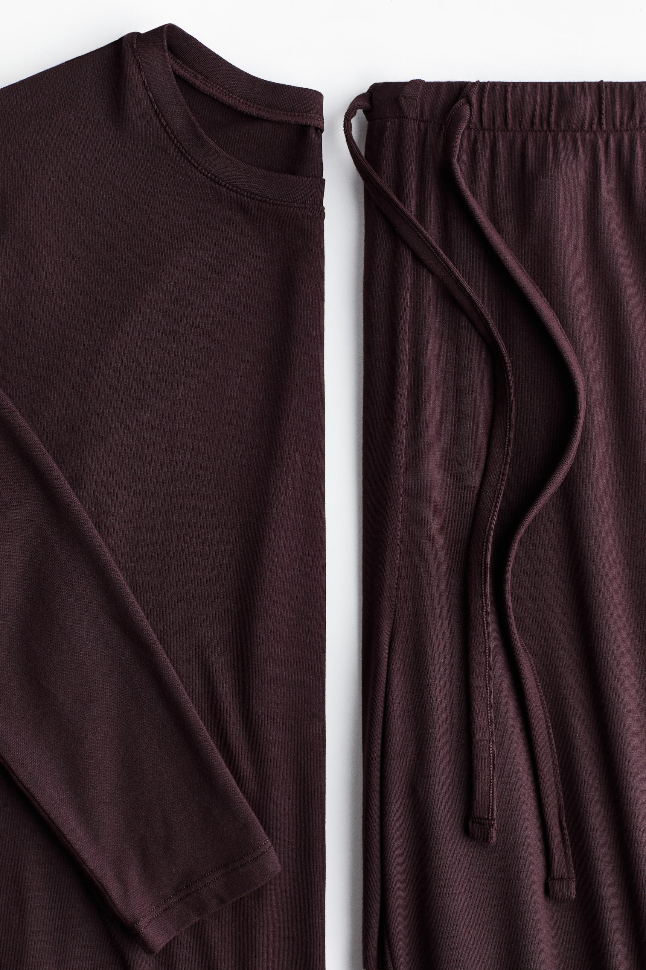 Grotere afbeelding bekijken: Een donker kastanjebruin tweedelig set ligt plat, met een lange mouwen top met ronde hals en bijpassende broek. De top toont een gladde stof met een gebonden halslijn, en de broek heeft een elastische tailleband met een trekkoord.