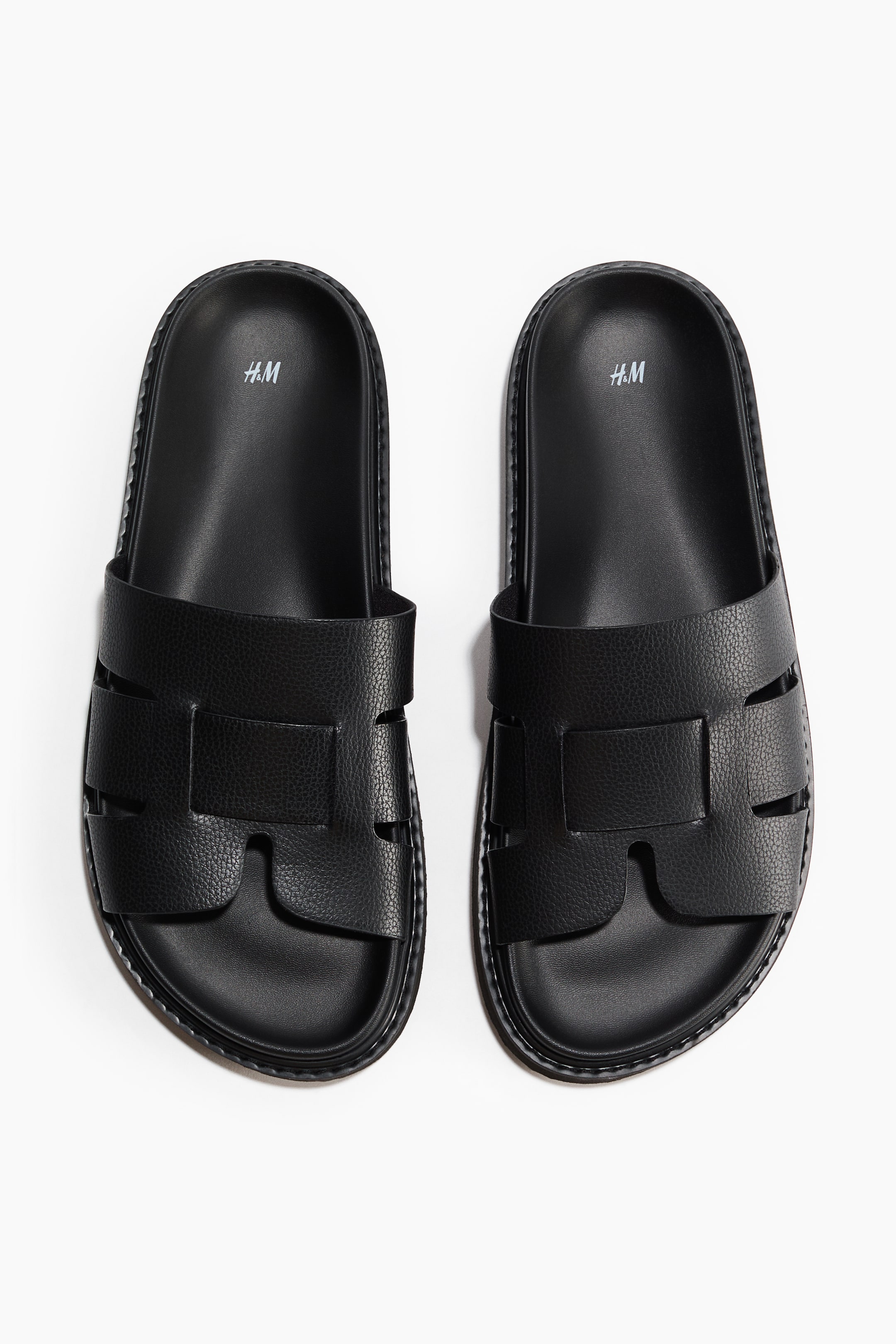 View larger image: A pair of black slide sandals is showcased from an overhead perspective. They boast moulded insoles adorned with a white 'H.M' logo and wide, textured straps crafted in a cut-out design. The sandals also feature moulded soles with an intricate pattern underneath.