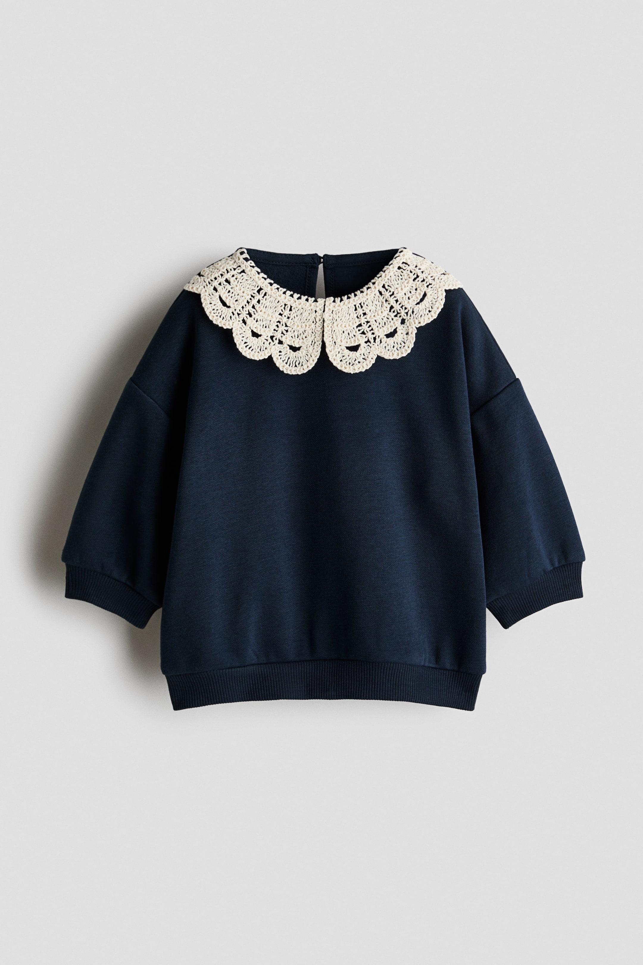 View larger image: A navy blue sweatshirt is shown from the front, featuring a large, cream-coloured, crocheted Peter Pan collar with scalloped details. The garment is designed with dropped shoulders, long sleeves with ribbed cuffs, and a matching ribbed hem.