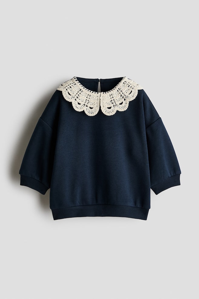 A navy blue sweatshirt is shown from the front, featuring a large, cream-coloured, crocheted Peter Pan collar with scalloped details. The garment is designed with dropped shoulders, long sleeves with ribbed cuffs, and a matching ribbed hem. - 1