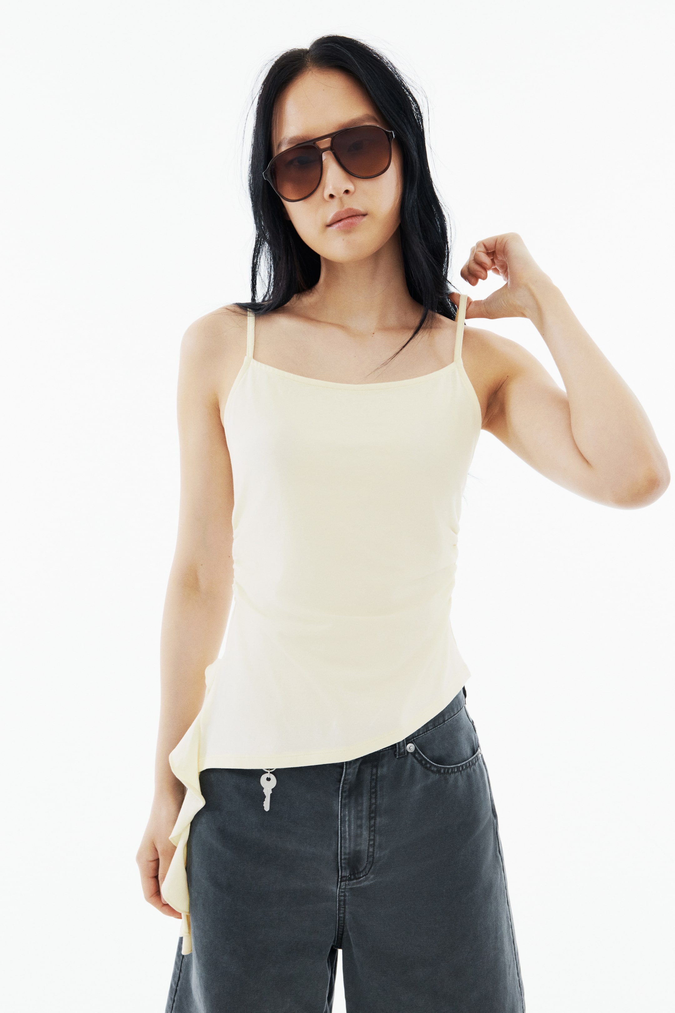View larger image: A light yellow camisole with thin straps and an asymmetrical hem with side ruching is being worn by the model, who is adjusting a shoulder strap while also wearing dark grey denim shorts with a key charm and dark brown sunglasses.