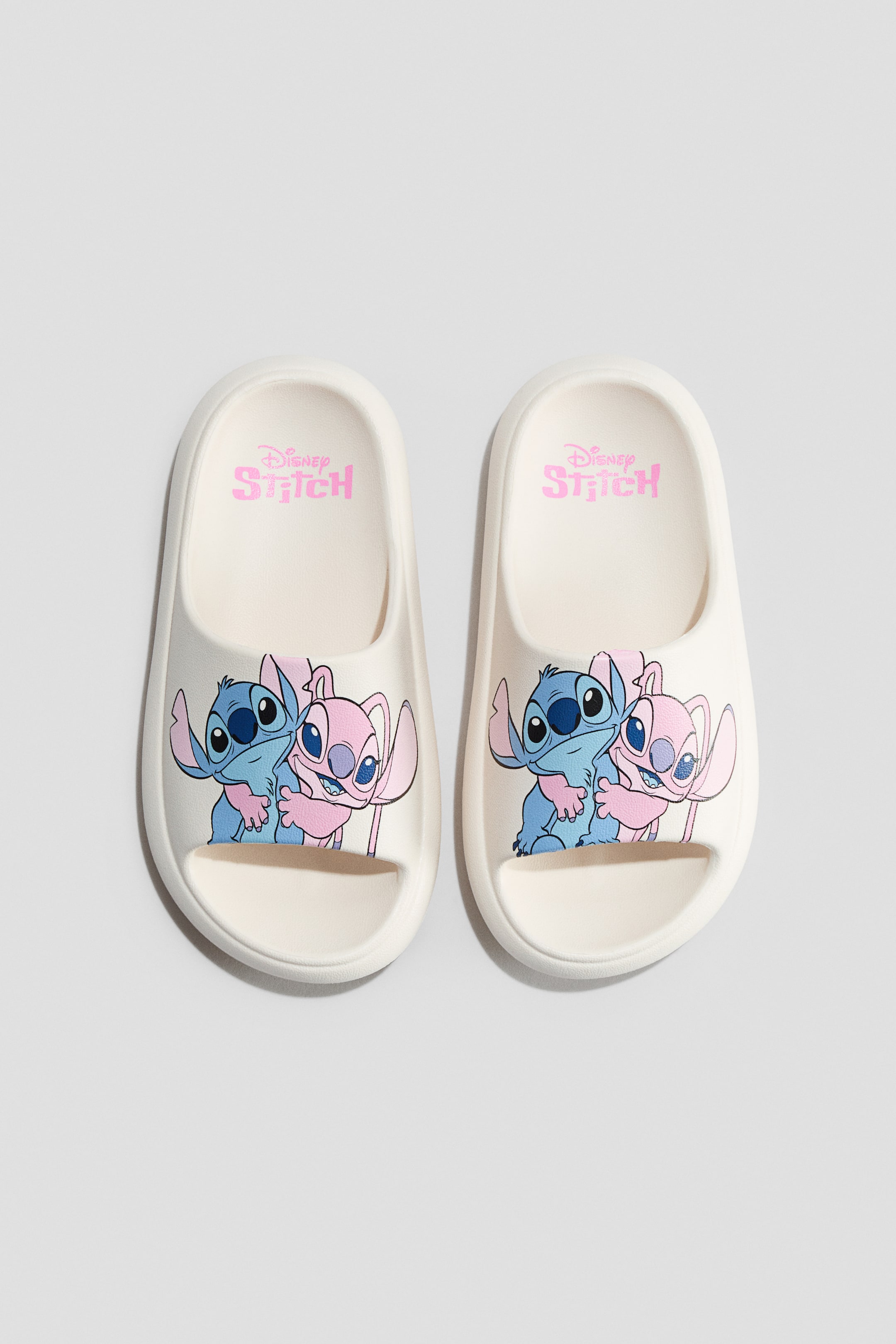 Se større billede: Formstøbte off-white poolsko viser et farverigt print af den blå karakter Stitch, der omfavner den lyserøde karakter Angel på hver skos forreste rem. Et lyserødt "Disney Stitch" logo ses på indersålen af hver sko.