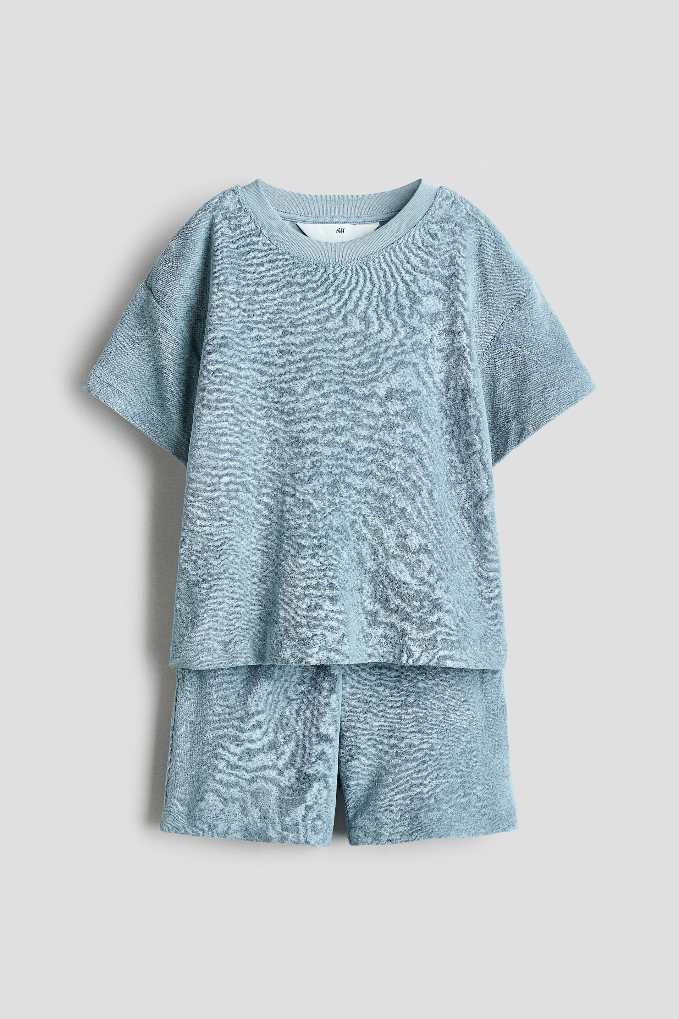 Agrandir l'image: Un ensemble en éponge bleu clair présente un t-shirt et un short, vus de face. Le t-shirt arbore une bordure en jersey côtelé autour de l'encolure et des épaules tombantes, tandis que le short affiche une taille élastiquée.
