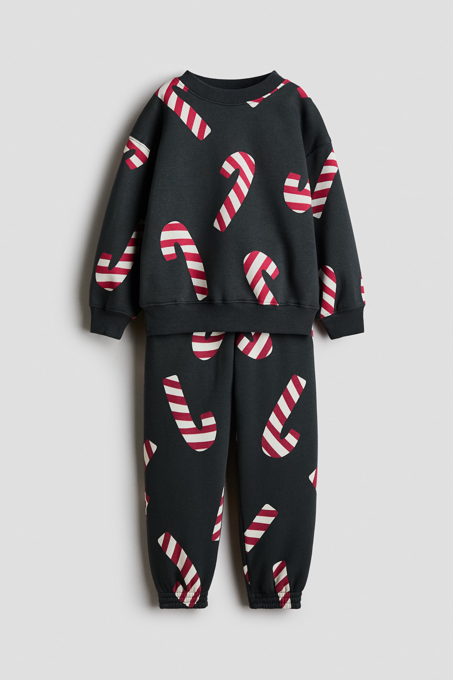 Eine Vorderansicht zeigt ein dunkelgraues zweiteiliges Fleece-Set bestehend aus einem langÃ¤rmeligen Sweatshirt und einer passenden Trainingshose, beide mit einem Allover-Print aus rot-weiÃ gestreiften Zuckerstangen. Das Sweatshirt prÃ¤sentiert sich mit geripptem Rundhalsausschnitt, Ã¼berschnittenen Schultern sowie gerippten BÃ¼ndchen und Saum, wÃ¤hrend die Hose mit elastischen BÃ¼ndchen abschlieÃt.