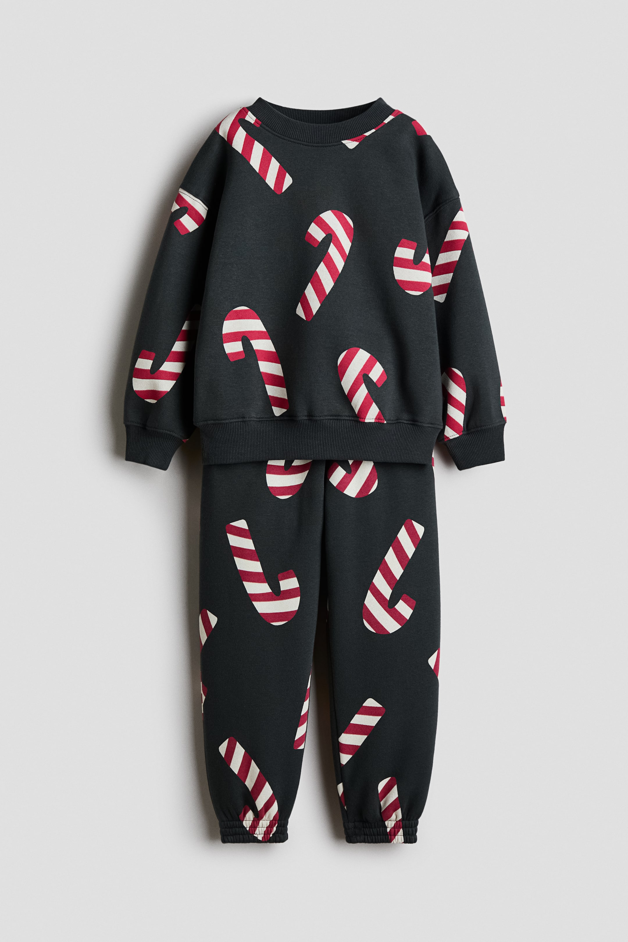 View larger image: A front view shows a dark grey two-piece fleece set consisting of a long-sleeve sweatshirt and matching track pants, both featuring an all-over print of red and white striped candy canes. The sweatshirt features a ribbed crew neck, dropped shoulders, and ribbed cuffs and hem, while the pants finish with elasticated cuffs.