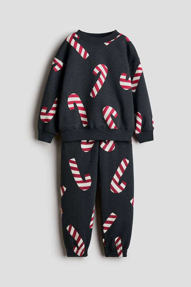 En frontvisning viser et mørkegrått todelt fleece-sett bestående av en langermet sweatshirt og matchende joggebukse, begge med et heldekkende trykk av røde og hvite stripete godteristenger. Sweatshirten har rund hals i ribbestrikk, droppede skuldre samt ribbestrikkede mansjetter og nederkant, mens buksen avsluttes med elastiske mansjetter. - 1
