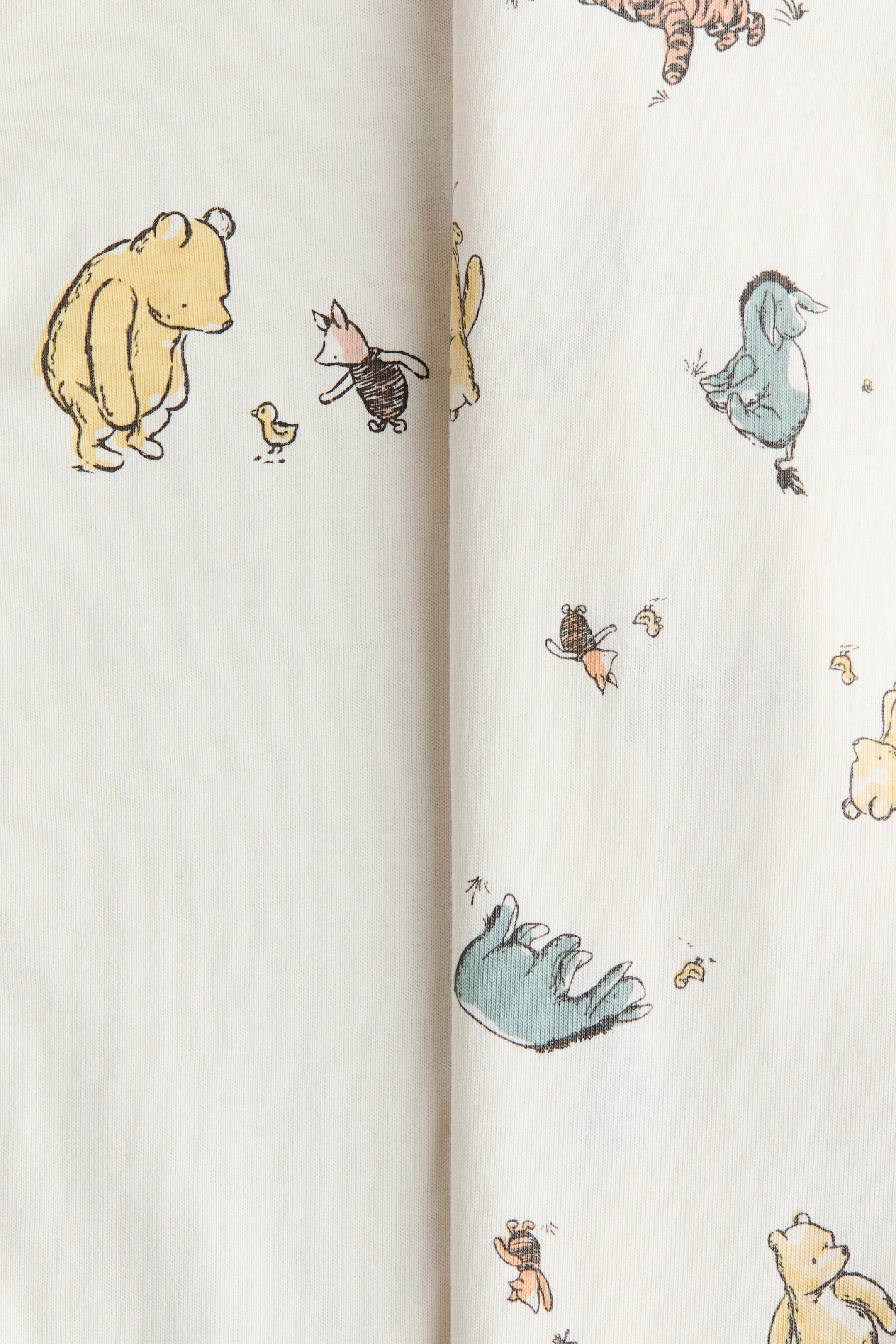 Visualizza l'immagine ingrandita: Una tutina realizzata in morbido jersey di cotone color crema presenta un motivo ripetuto di personaggi di Winnie the Pooh, tra cui Winnie, Pimpi, Ih-Oh e Tigro, sparsi sul tessuto.