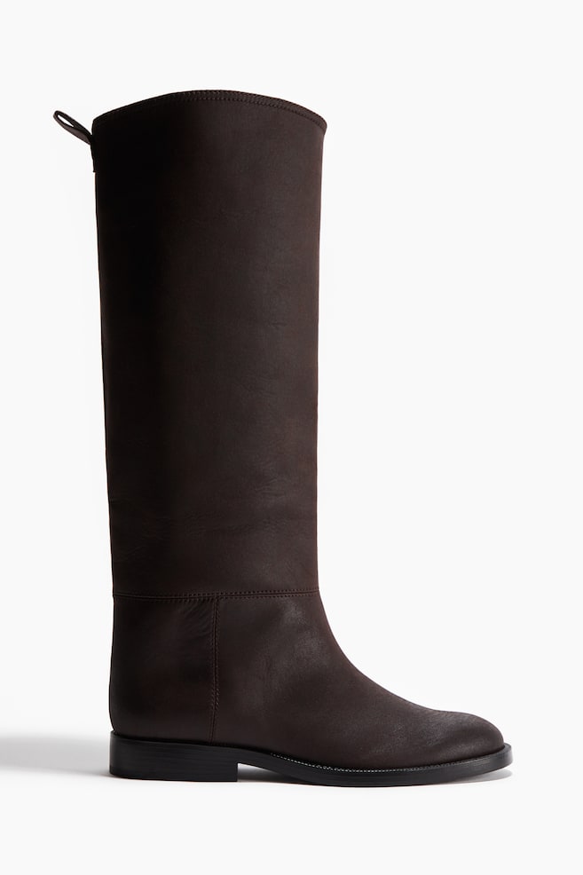 A single dark brown knee-high boot is shown in a side view, featuring a smooth, matte finish, a rounded toe, and a low, flat black heel. A horizontal seam is visible above the ankle, and a small pull tab is at the top back of the shaft. - 1