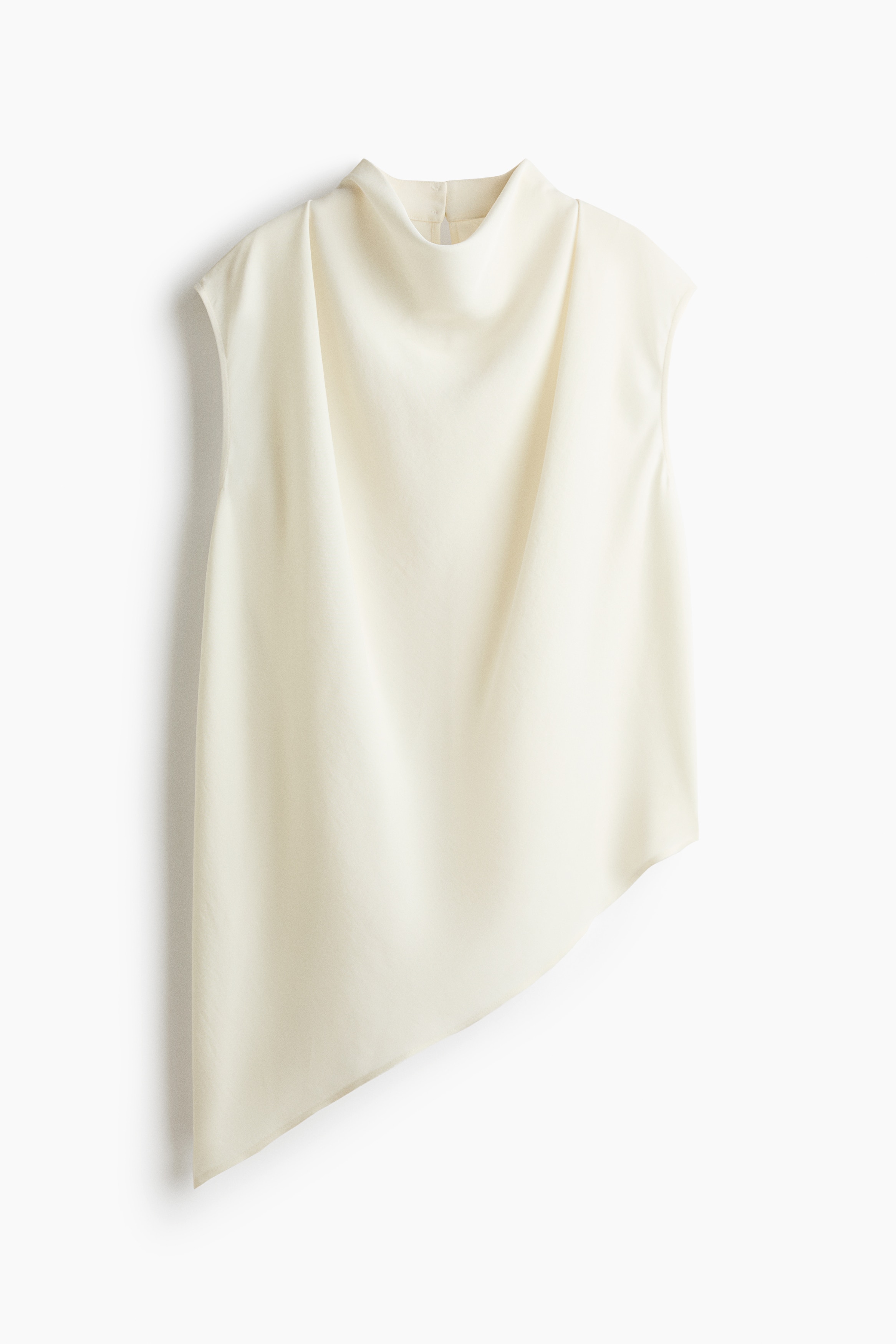 A cream-coloured sleeveless top featuring a cowl neckline, pleated shoulders, and an asymmetrical hem is worn by the model. The top is styled with light-wash, wide-leg blue jeans.