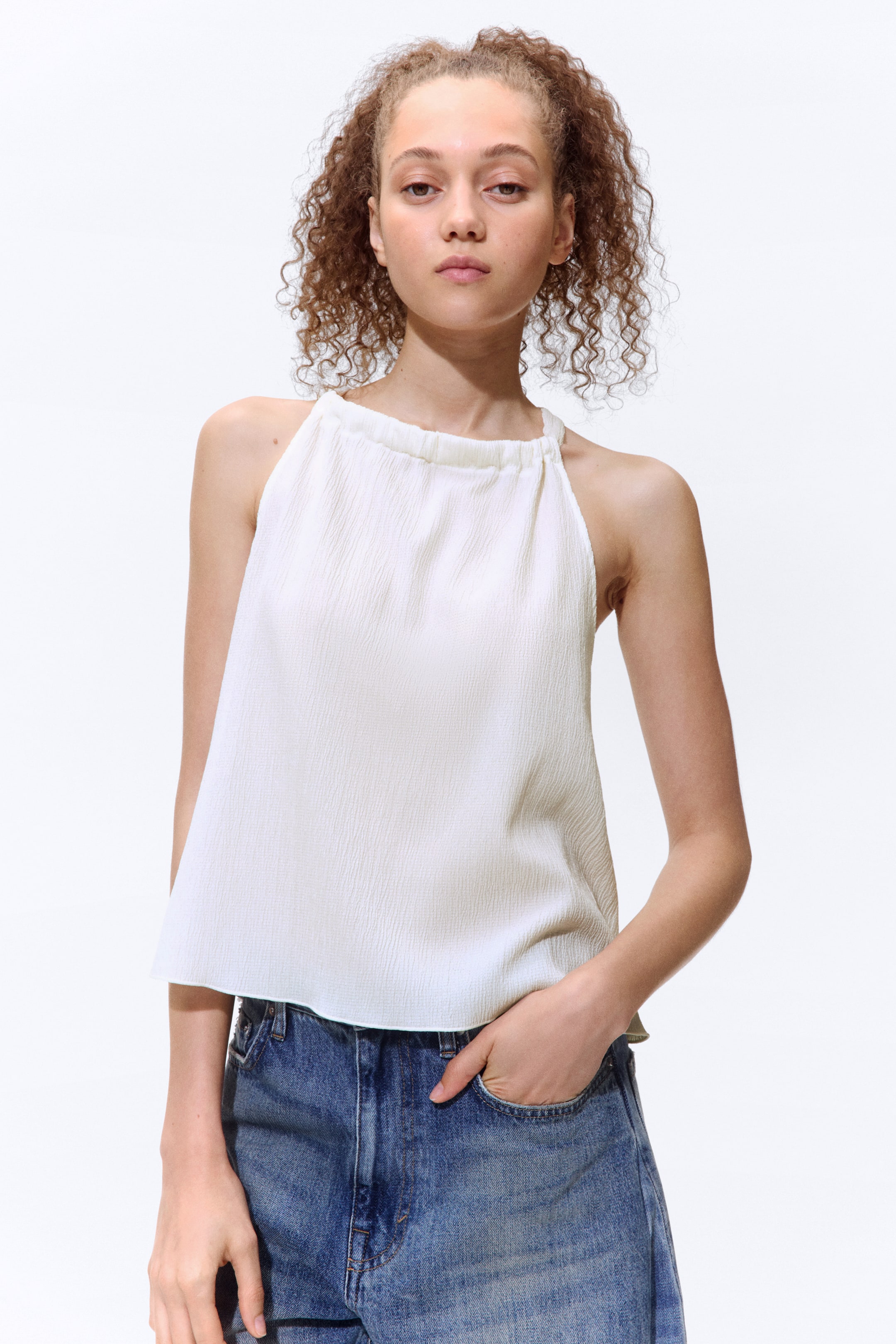 View larger image: The model is wearing a short, sleeveless top crafted from crinkled off-white jersey. It showcases a round neckline with a wide drawstring that creates gathering, a narrow cut at the shoulders, and an overlocked hem, styled with blue jeans.