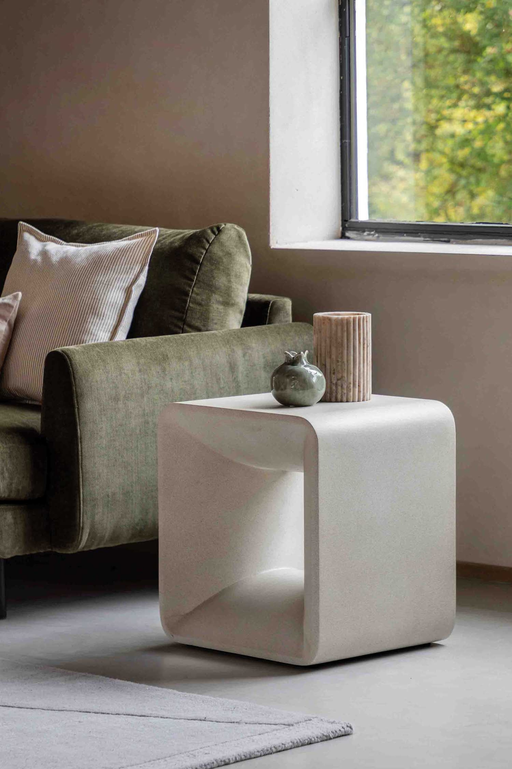 View larger image: A light-toned, C-shaped side table with rounded edges and a textured matte finish is positioned, angled slightly to the right, showcasing its open interior. A small green decorative object and a ribbed cylindrical object are resting on its top surface.