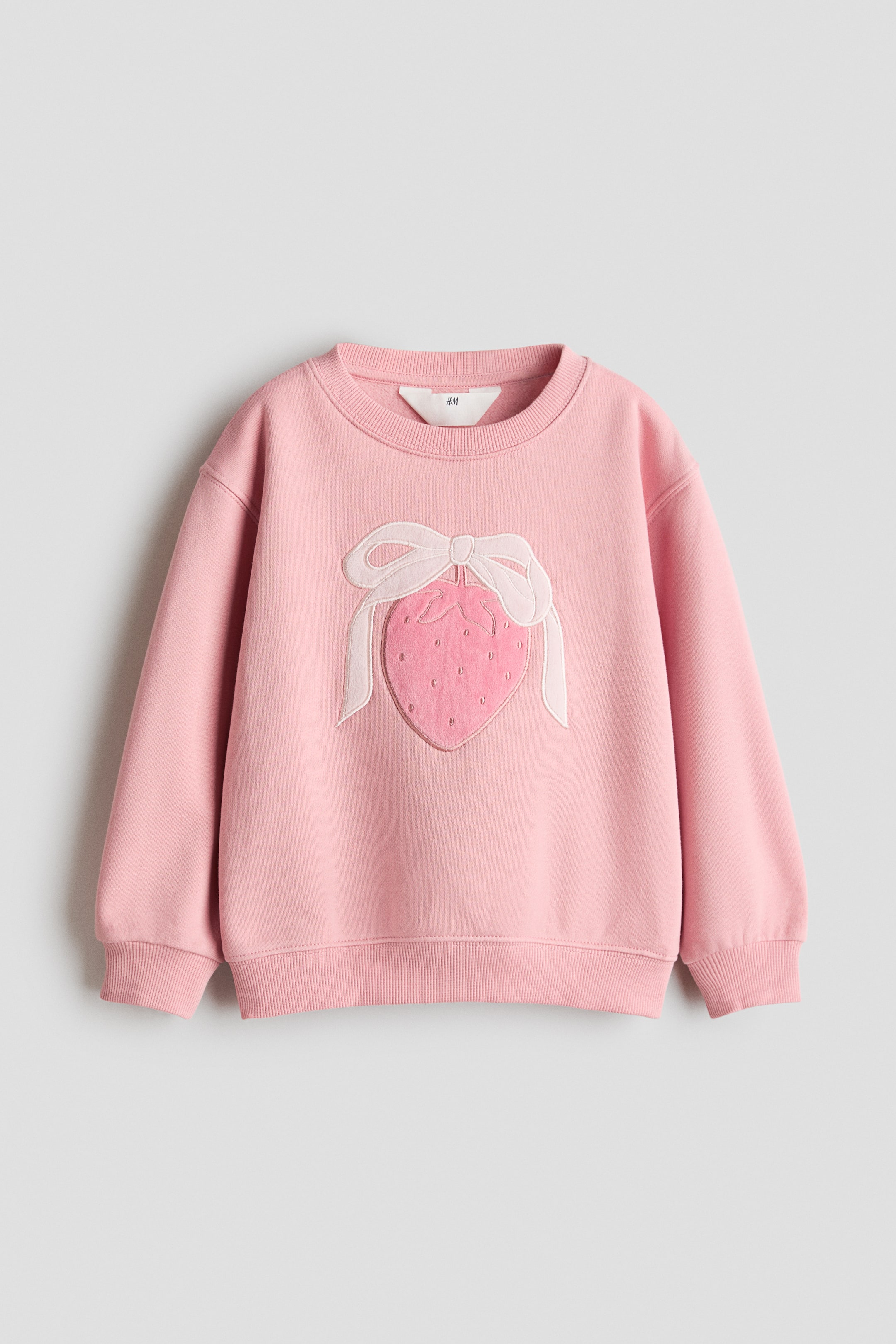 View larger image: A pink jumper is showcased from the front, highlighting a large textured strawberry motif with a light pink bow on the chest. It features a round neckline, gently dropped long sleeves, and ribbing at the neckline, cuffs, and hem.