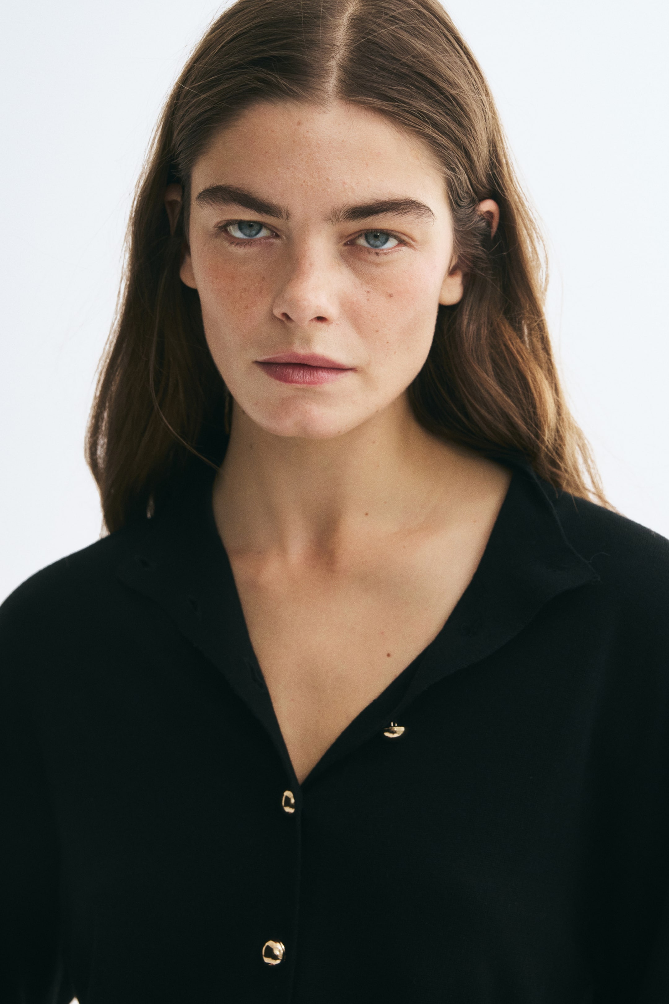 Größeres Bild ansehen: Die Model trägt eine schwarze Strickjacke mit weichem Kragen und kleinen goldfarbenen Knöpfen auf der Vorderseite. Die oberen Knöpfe sind offen getragen, um einen V-Ausschnitt zu erzeugen.
