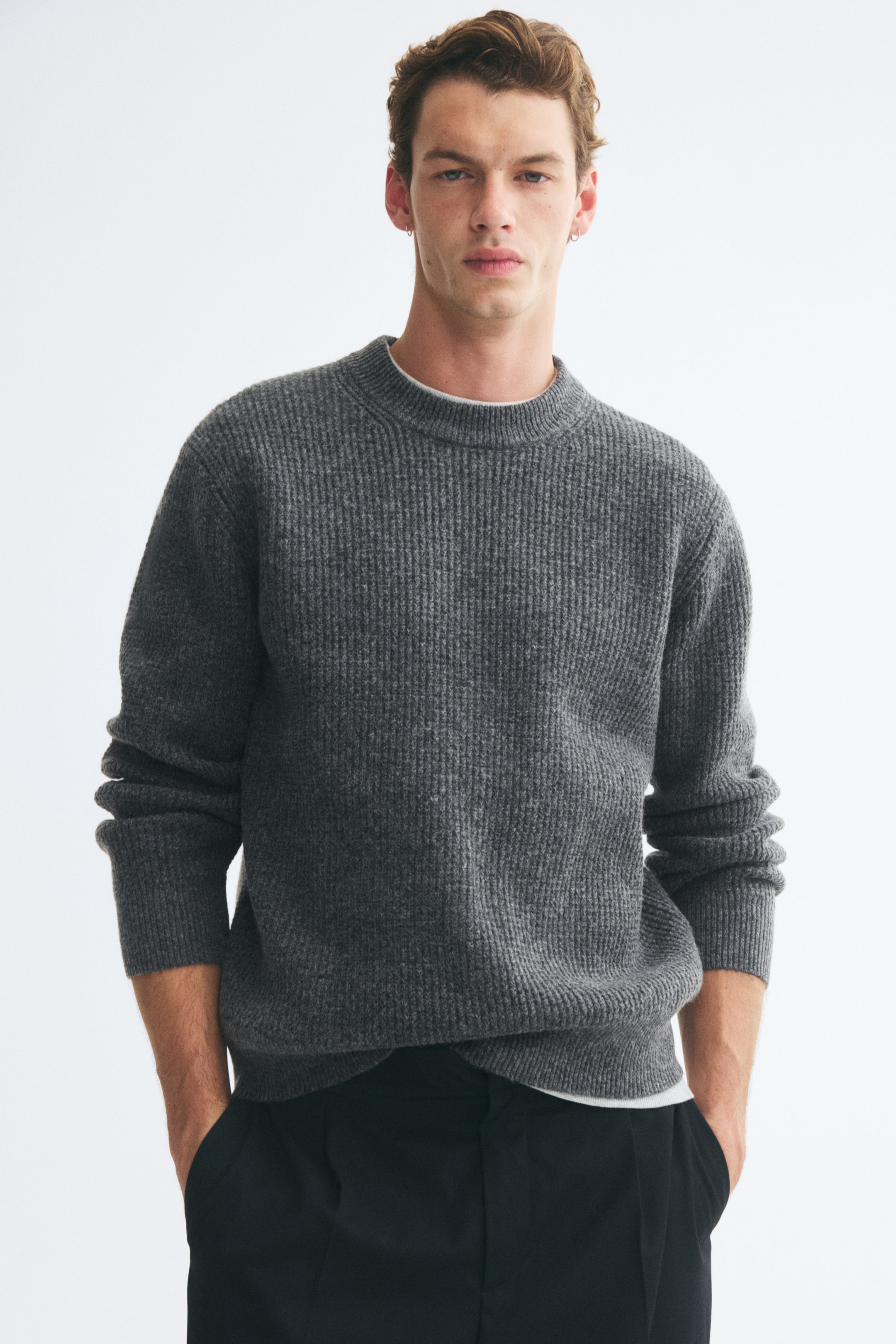Agrandir l'image: Le mannequin porte un pull gris en maille texturée, avec les mains dans les poches d’un pantalon noir. Le pull présente une coupe décontractée, un col rond côtelé porté sur un t-shirt blanc, ainsi que des finitions côtelées aux poignets et à l’ourlet.
