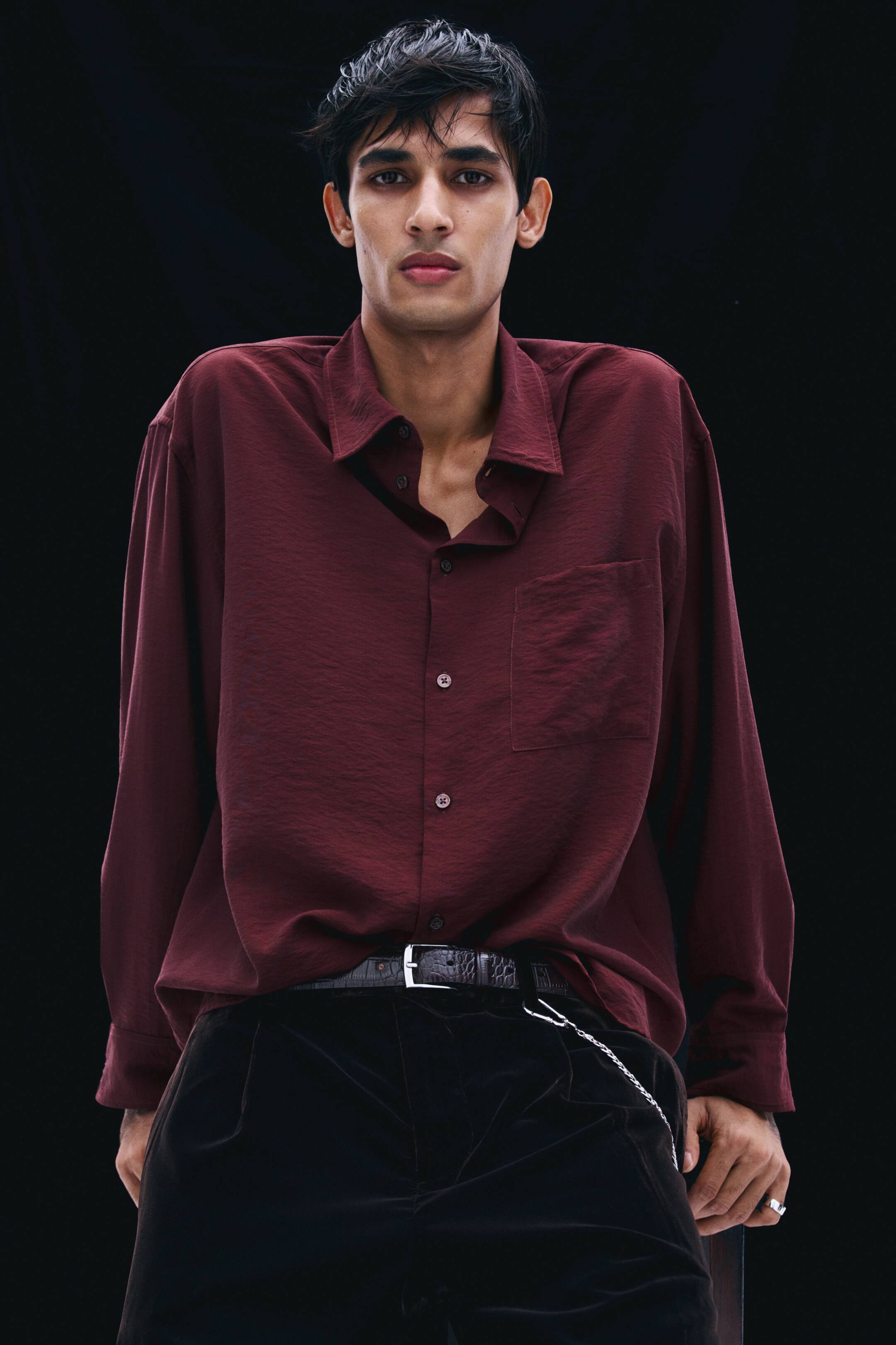 View larger image: A burgundy long-sleeve shirt in a lightly crinkled fabric is worn by the model, tucked into dark trousers with a black belt. The relaxed-fit shirt features a collar, a partially unbuttoned front placket, and a single patch chest pocket.