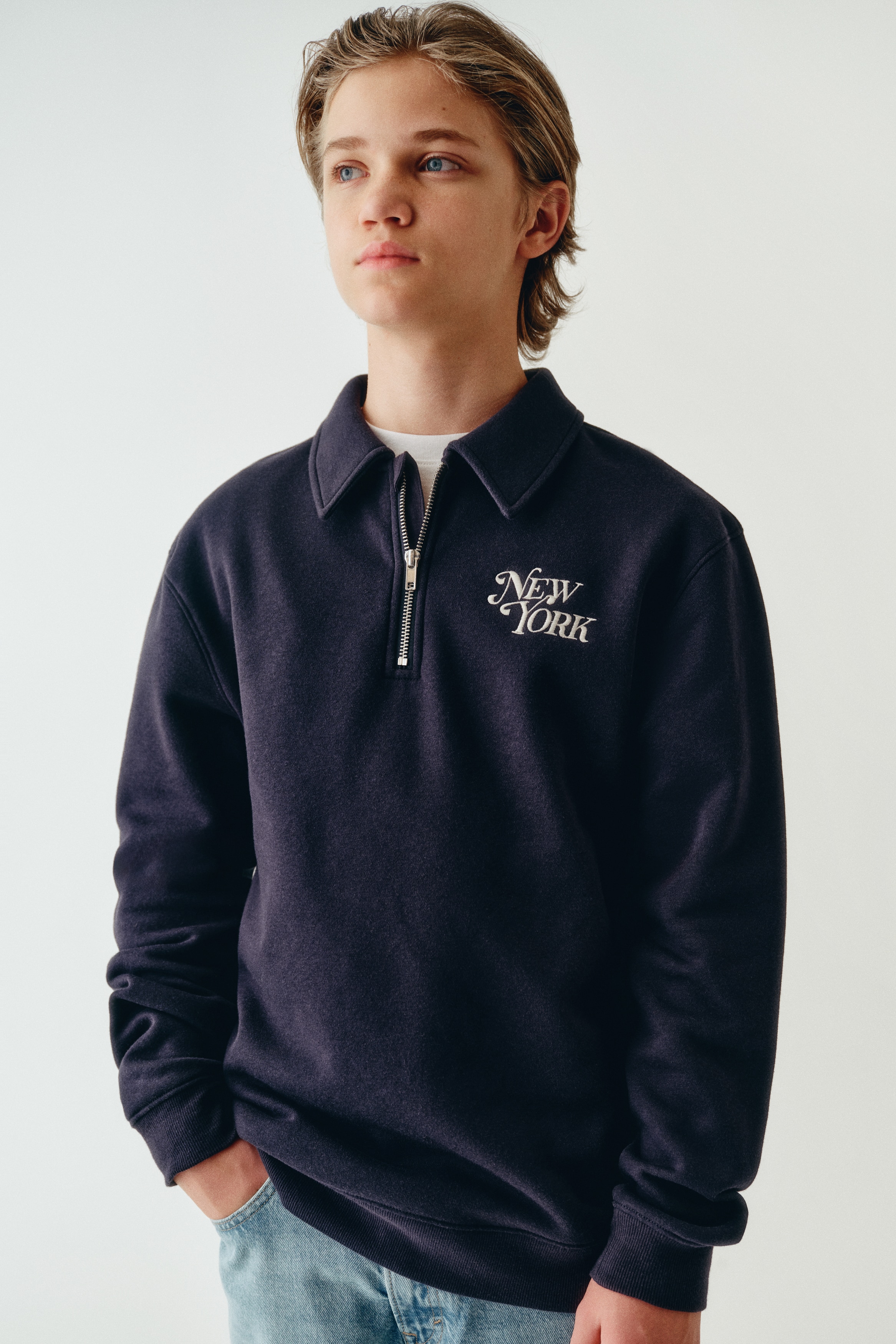 Má»t chiáº¿c Ã¡o sweatshirt ná»­a khÃ³a mÃ u xanh navy Äang ÄÆ°á»£c chá»¥p chÃ­nh diá»n, ná»i báº­t vá»i cá» polo cÃ³ phom Äá»©ng, khÃ³a kÃ©o kim loáº¡i tÃ´ng báº¡c, vÃ  dÃ²ng chá»¯ "New York" ÄÆ°á»£c thÃªu báº±ng chá»¯ viáº¿t tay mÃ u tráº¯ng á» ngá»±c trÃ¡i. Thiáº¿t káº¿ thá» hiá»n tay dÃ i vá»i bo tay dá»t gÃ¢n vÃ  lai Ã¡o dá»t gÃ¢n Äá»ng bá».