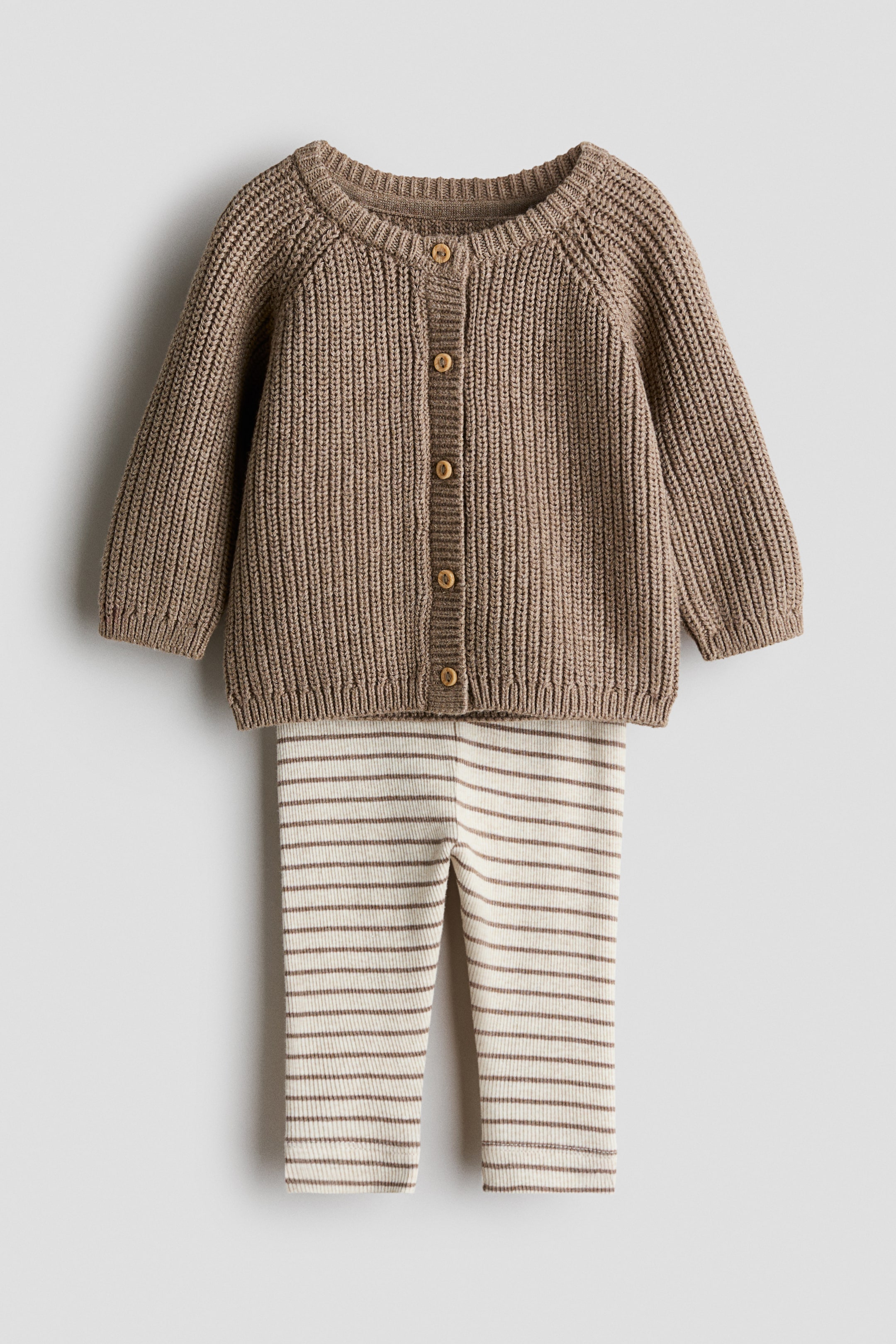 Grösseres Bild ansehen: Ein zweiteiliges Baby-Outfit, von vorne betrachtet, besteht aus einer braunen, gerippten Strickjacke mit rundem Ausschnitt und fünf runden, braunen Knöpfen an der vorderen Knopfleiste, zusammen mit cremefarbenen, gerippten Strickleggings mit dünnen, horizontalen, braunen Streifen.