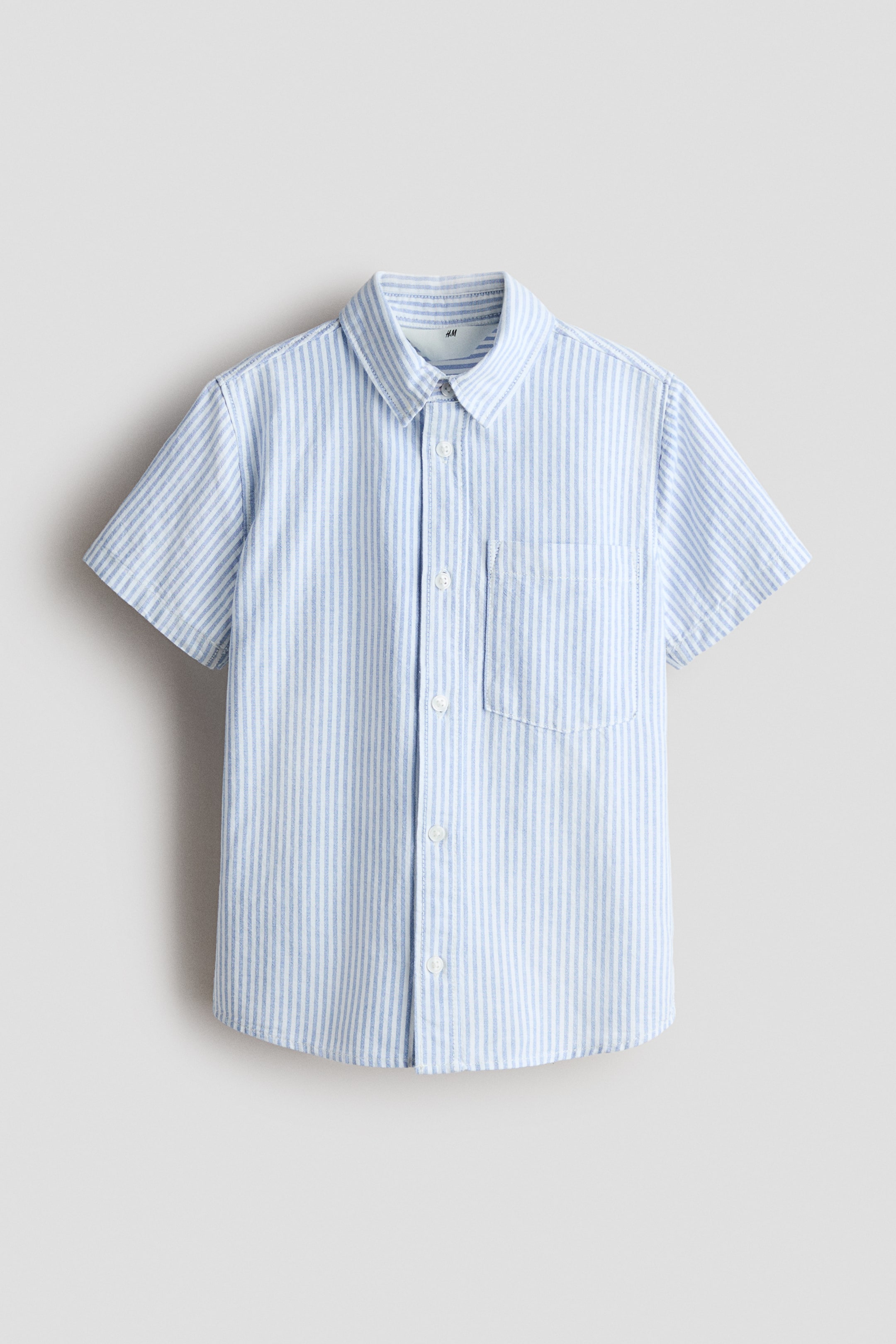 View larger image: The short-sleeve shirt is shown from the front, showcasing a light blue and white striped cotton fabric. It features a turn-down collar, buttons along the front, a single chest pocket on the left side, and a softly rounded hem.