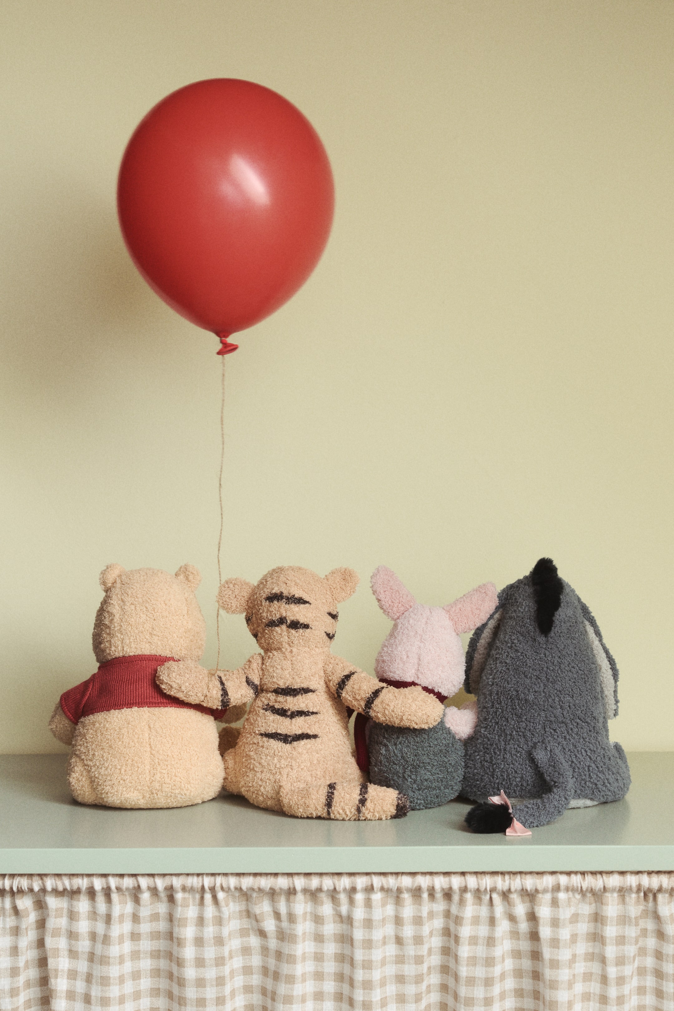 拡大画像を表示: テディ生地で作られた4つのぬいぐるみは、刺繍のディテールが施され、100エーカーの森のキャラクターとしてデザインされています。これらのぬいぐるみは、背中を向けて座っています。薄茶色のクマは赤いニットのトップを着ており、赤い風船の紐を持っています。その隣には、縞模様のあるタン色のトラ、ピンクのブタ、そして尾にピンクのリボンをつけた灰色のロバがいます。