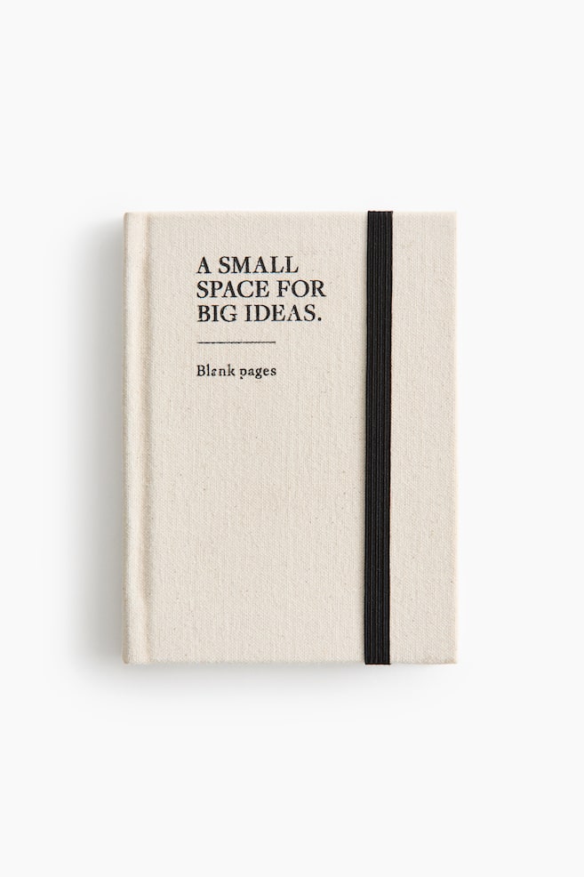 A small notebook featuring an off-white cotton cover is lying flat in a front view, showcasing the black printed text âA SMALL SPACE FOR BIG IDEAS.â and âBlank pages.â A thick black elastic band runs vertically along the right side to secure the cover. - 1