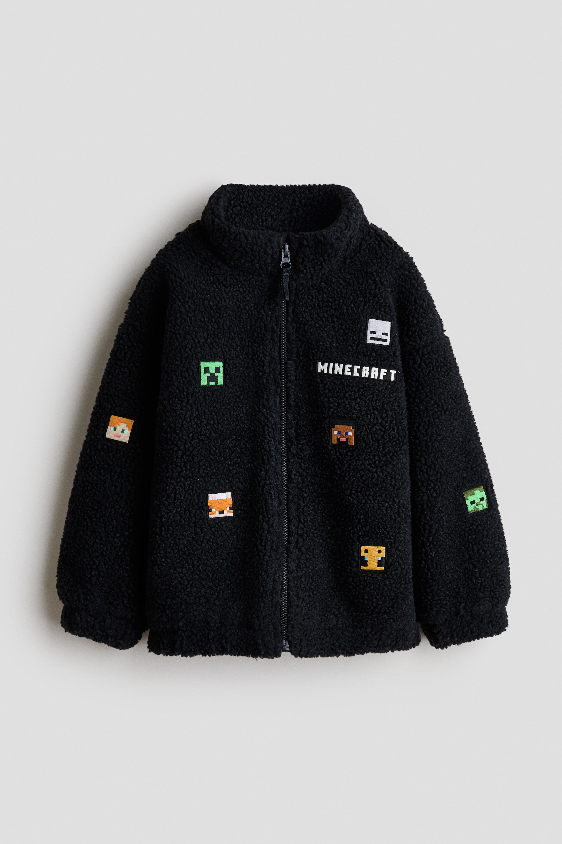 Agrandir l'image: Une veste en polaire sherpa noire est présentée de face, dotée d'une fermeture éclair intégrale et d'un col montant. Elle est ornée de plusieurs petits patchs brodés pixelisés représentant des personnages et des objets de Minecraft, ainsi que "MINECRAFT" brodé en texte blanc sur la poitrine gauche.