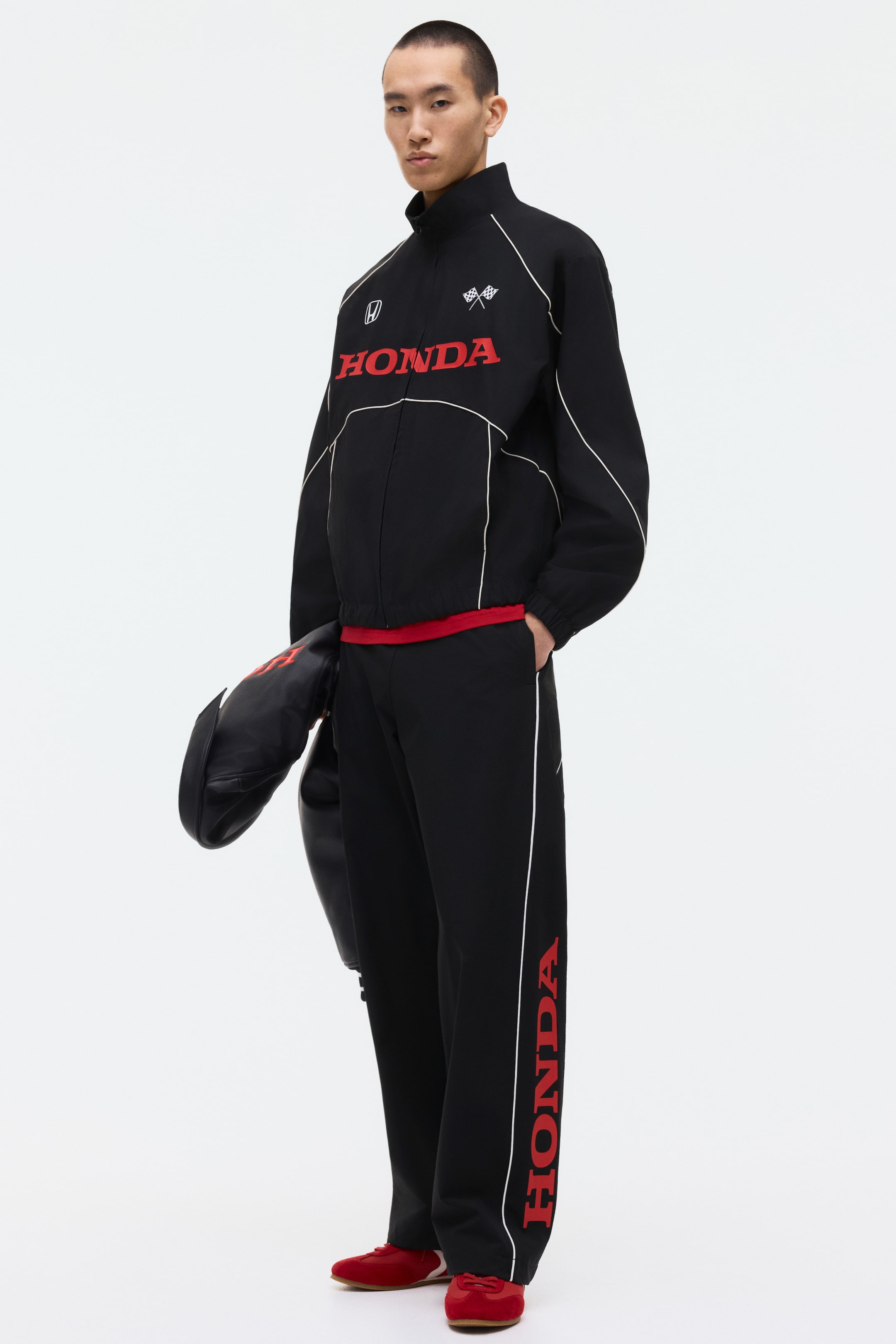 View larger image: These black track pants feature a front-facing design with a covered elastic waistband, welt side pockets, and white piping along the outer seams of both legs. A red "HONDA" print runs vertically down the outer side of the right leg, all in a relaxed fit.