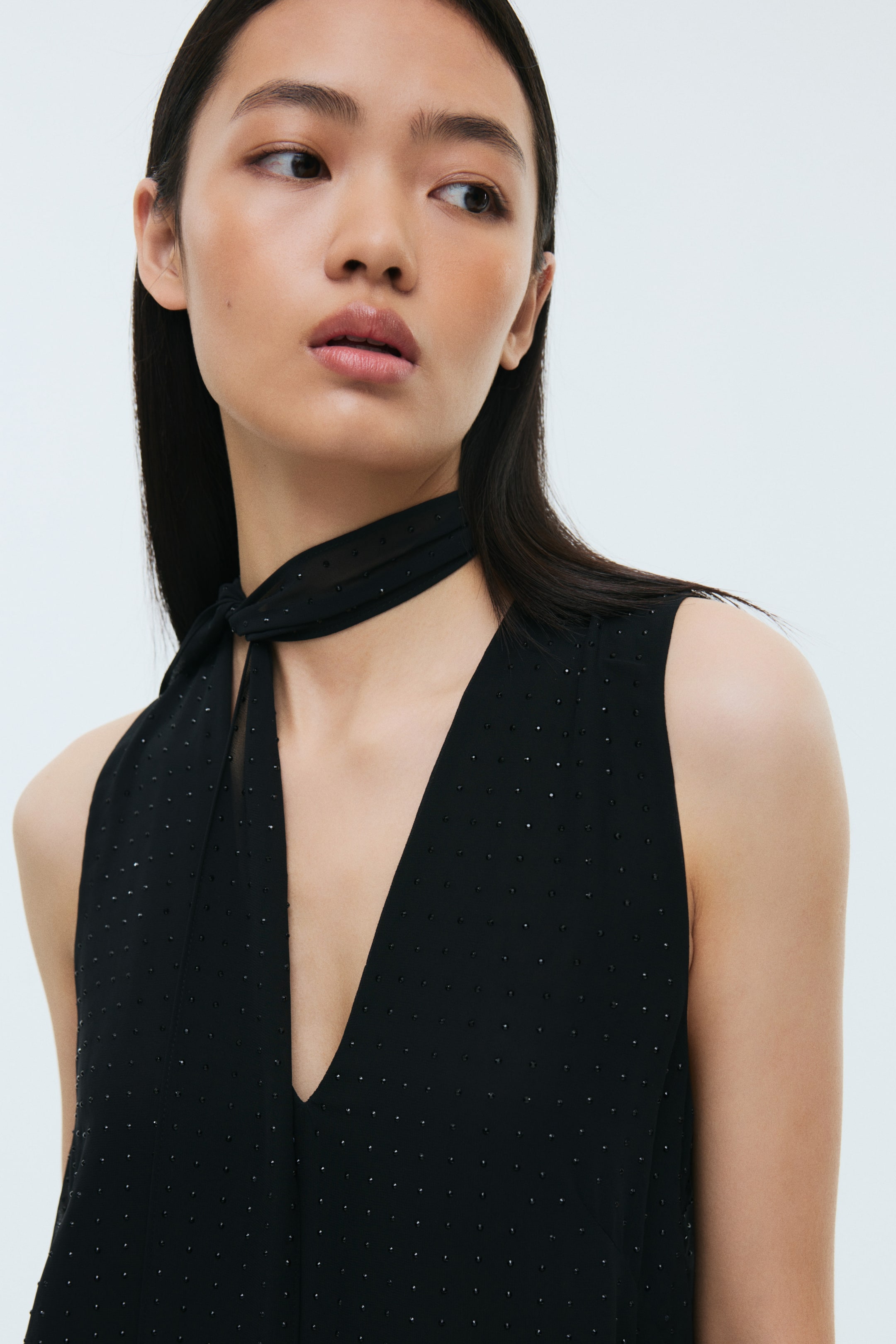 Ampliar la imagen: El producto es una blusa negra sin mangas, vista desde el frente, que presenta un escote profundo en V. Pequeños puntos brillantes están dispersos por el tejido, y una bufanda estrecha a juego está atada alrededor del cuello.