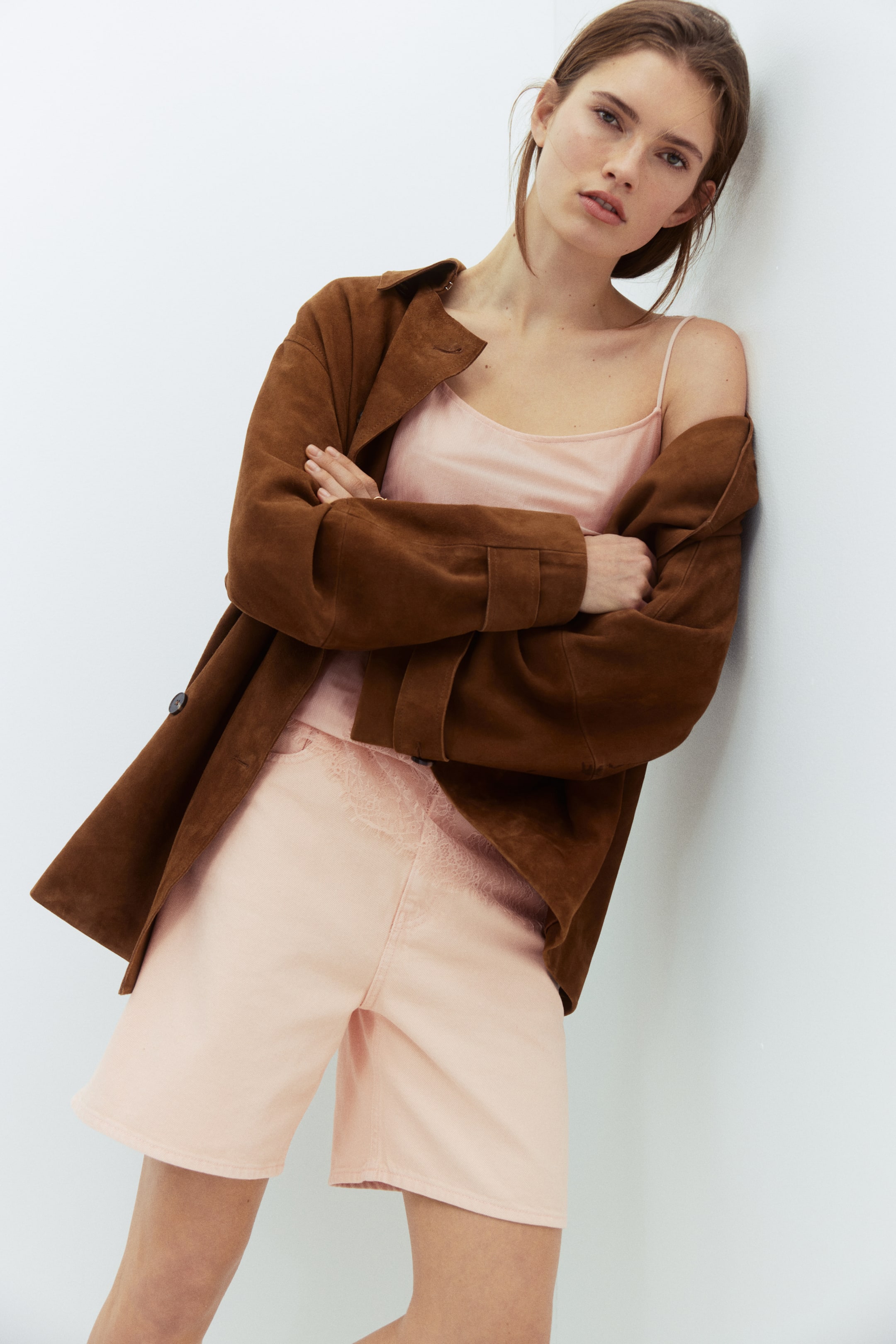 View larger image: A brown suede jacket is worn open, its sleeves pushed up to reveal strap detailing at the cuffs. It is layered over a light pink camisole and matching high-waisted shorts, both featuring lace detailing along the waistline.