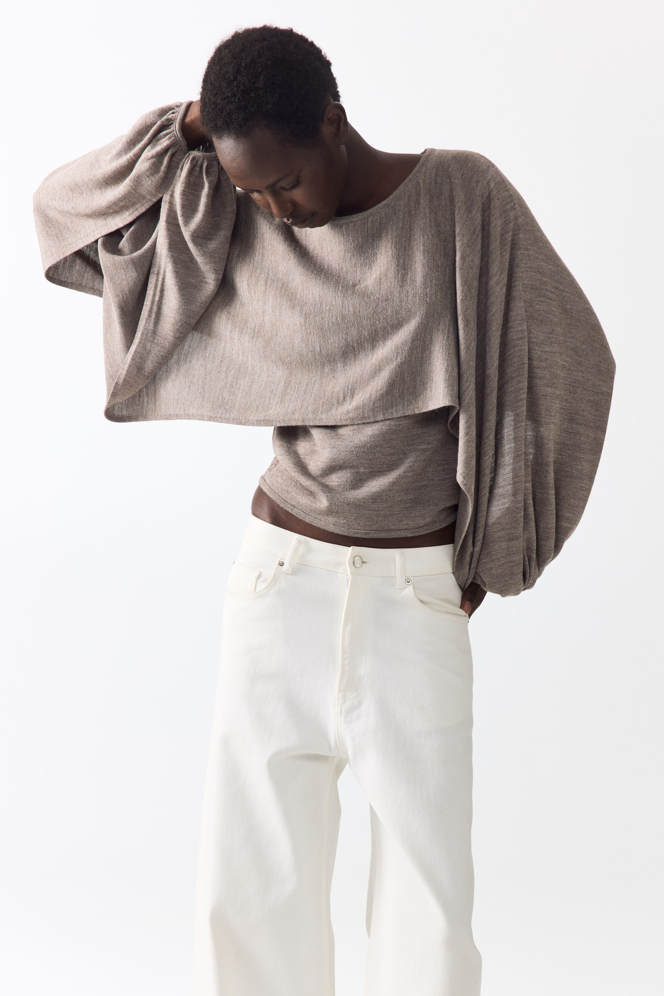 Grotere afbeelding bekijken: Een taupe gebreide top met een brede halslijn, volumineuze ballonmouwen met aansluitende manchetten en een gelaagd ontwerp met een kortere overlay. De aansluitende onderlaag van de top wordt in hoog getailleerde witte wijde broekspijpen gestopt.