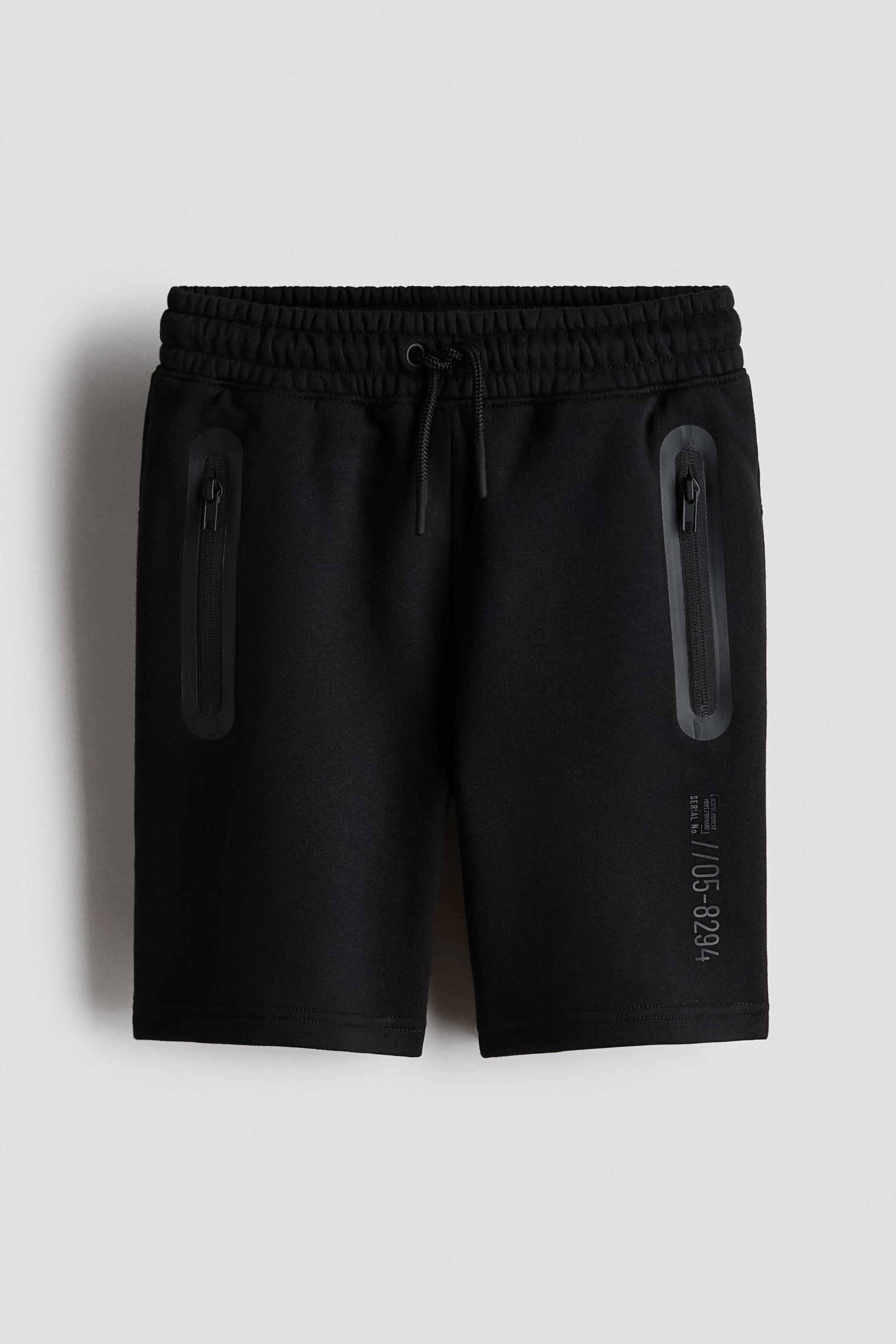 Se større billede: Sorte shorts har en elastisk linning med snor samt to lodrette lynlåslommer med forseglede kanter foran. Et gråt serienummerprint ses på nederste del af højre ben.